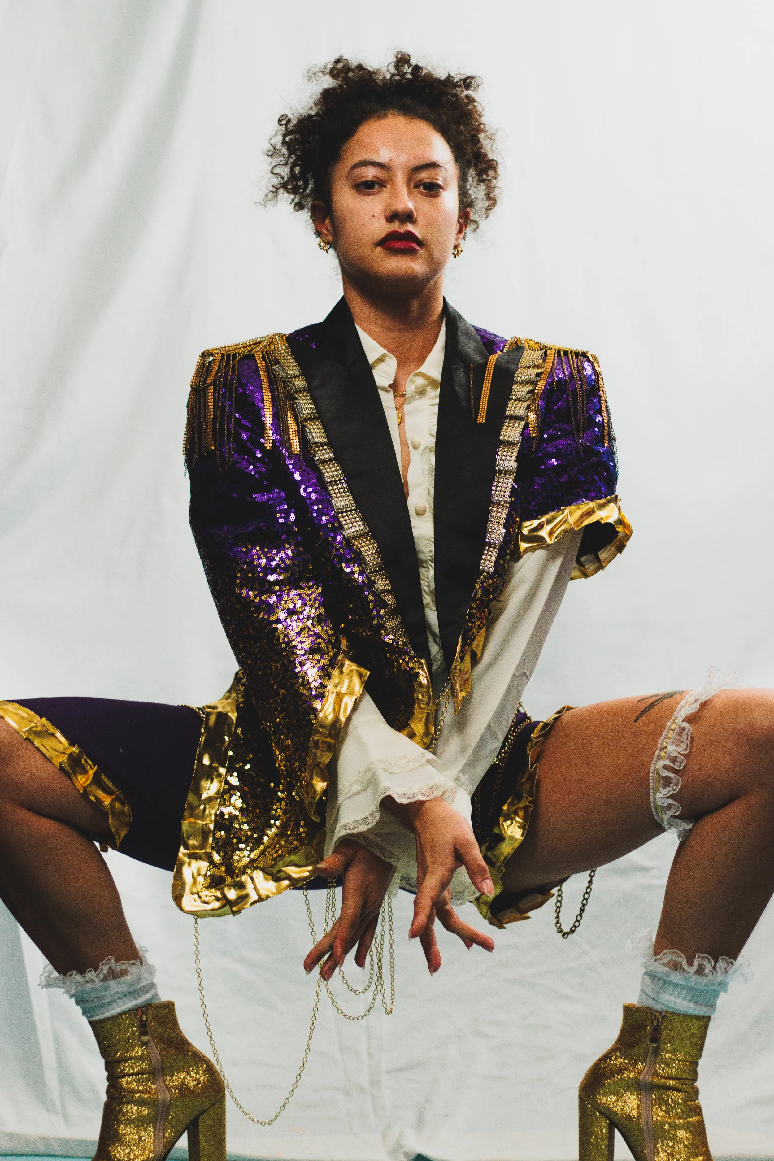 Portrait of a person in a purple-and-gold sequined military-style jacket, white ruffled shirt, and gold glitter boots, seated against a white backdrop.