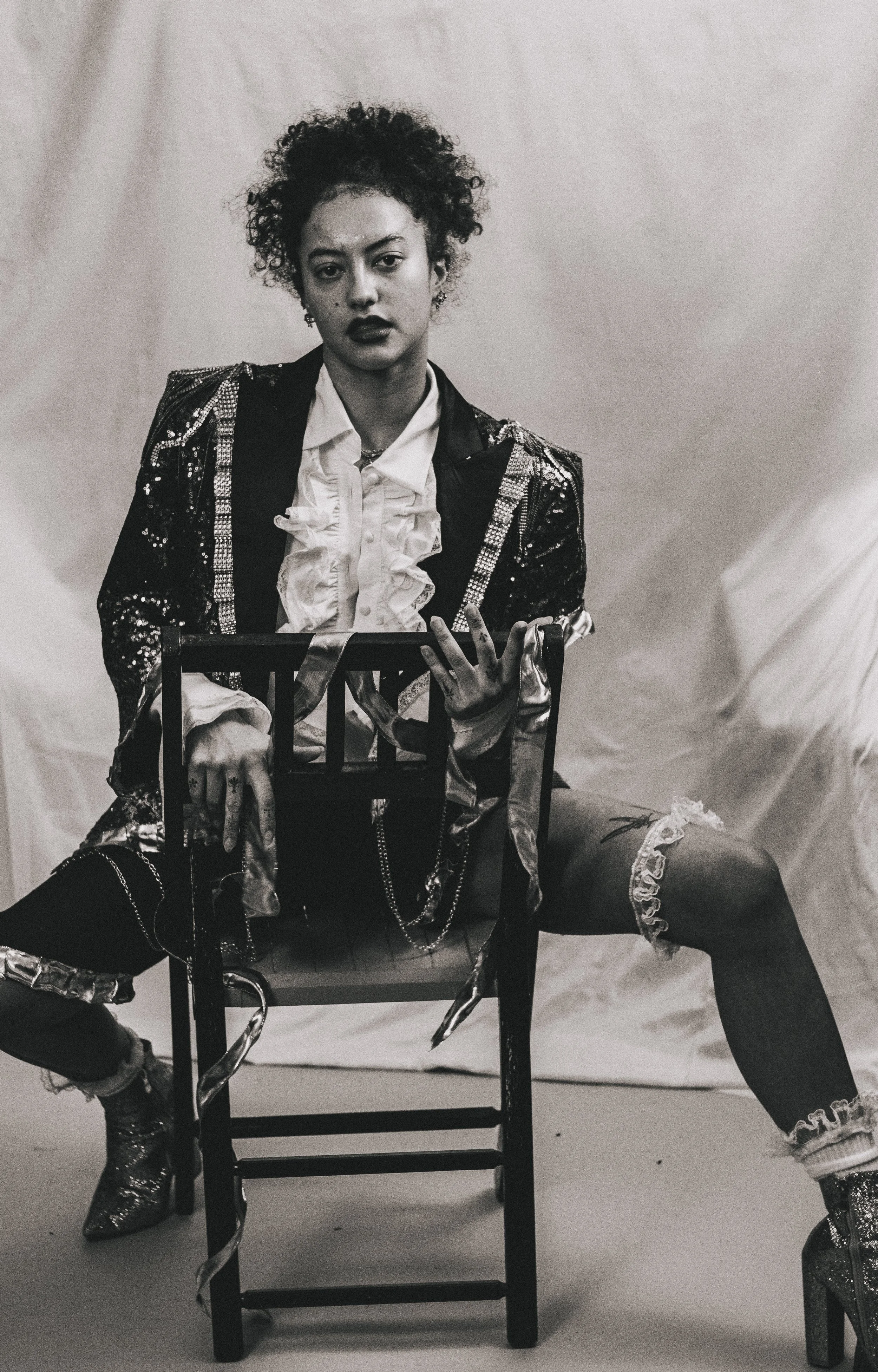 Black-and-white portrait of a curly-haired person in a sequined jacket and ruffled shirt sitting behind a wooden chair.