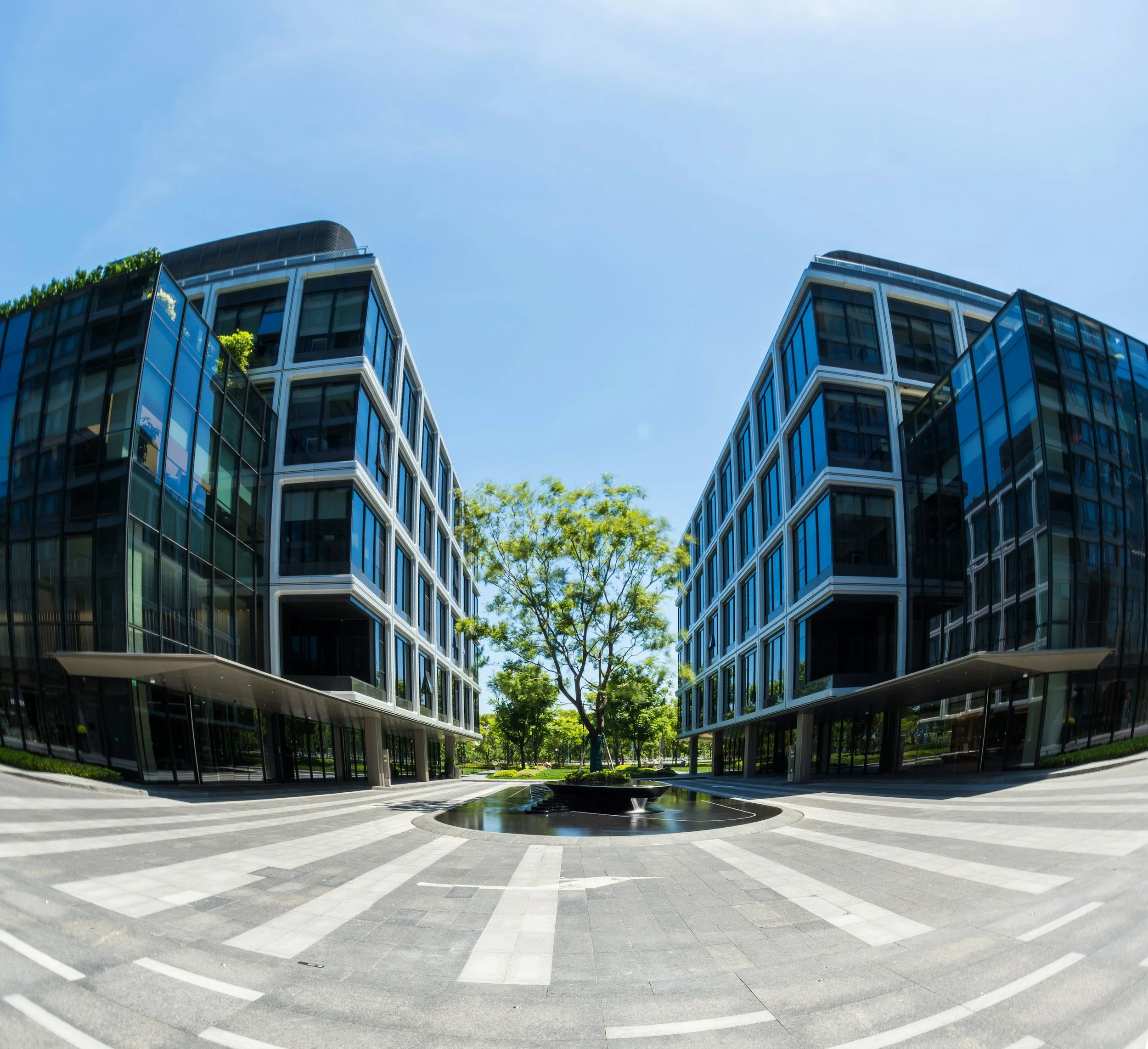 Modern glass buildings with reflective windows, a small fountain, and a paved circular plaza with white striped patterns, surrounded by green trees under a blue sky.