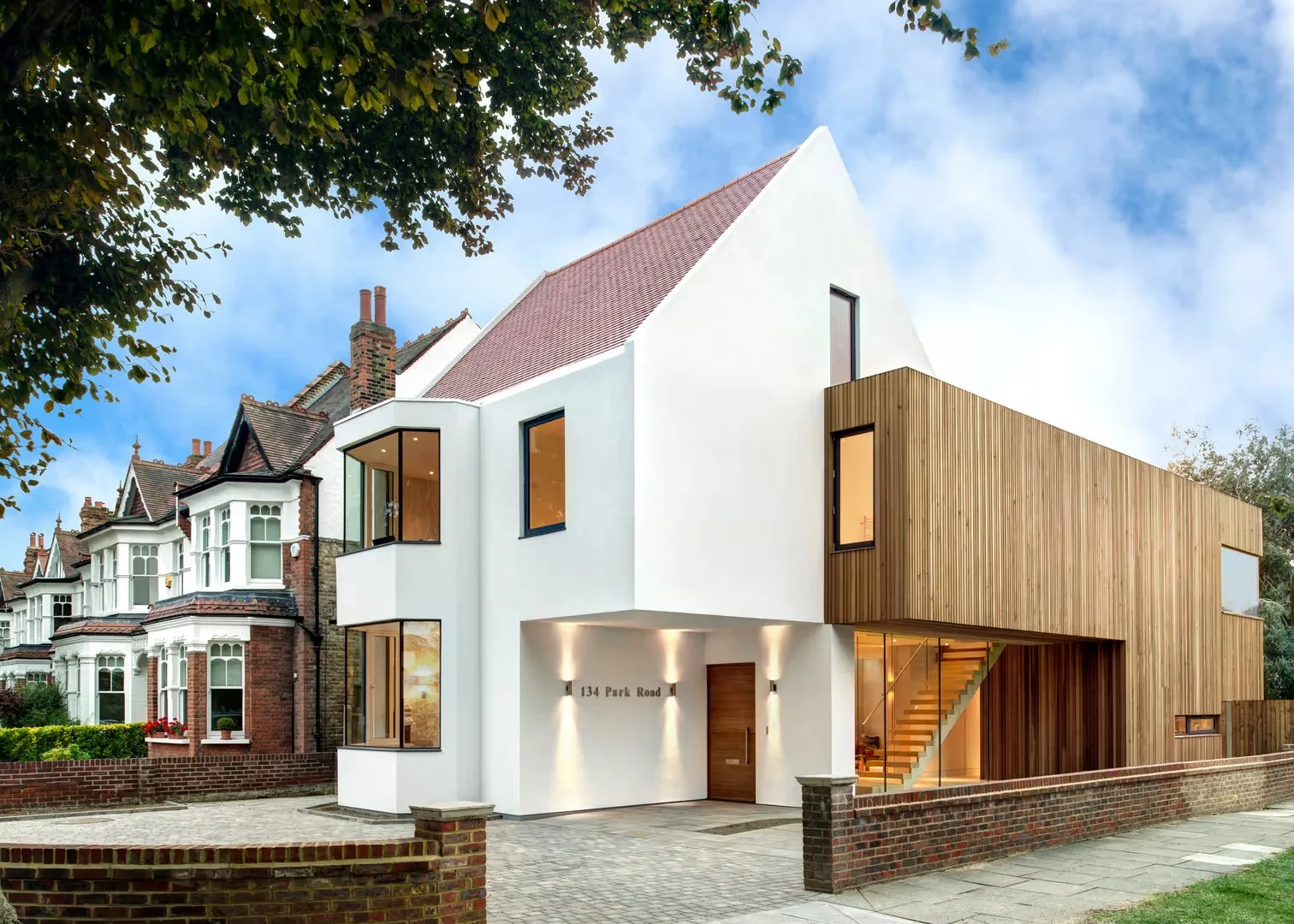 Modern house with white and wooden exterior, large windows, and a staircase visible through a glass wall, located at 134 Park Road, with neighboring traditional brick houses and trees.