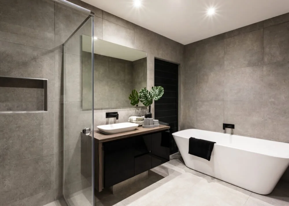 Modern bathroom with gray tiled walls, a white bathtub with a black towel, a black vanity with a vessel sink, a large mirror, a plant, and recessed ceiling lights.