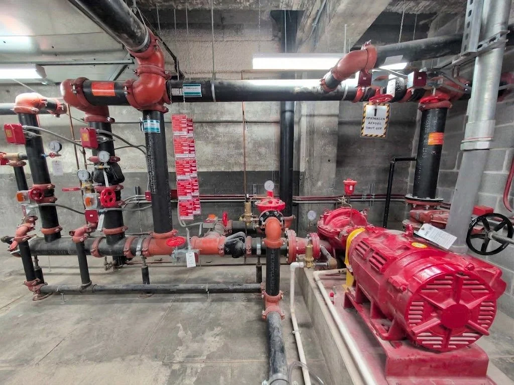 A network of red and black pipes, valves, and gauges in an industrial or mechanical room, with a red pump and pressure gauges attached.