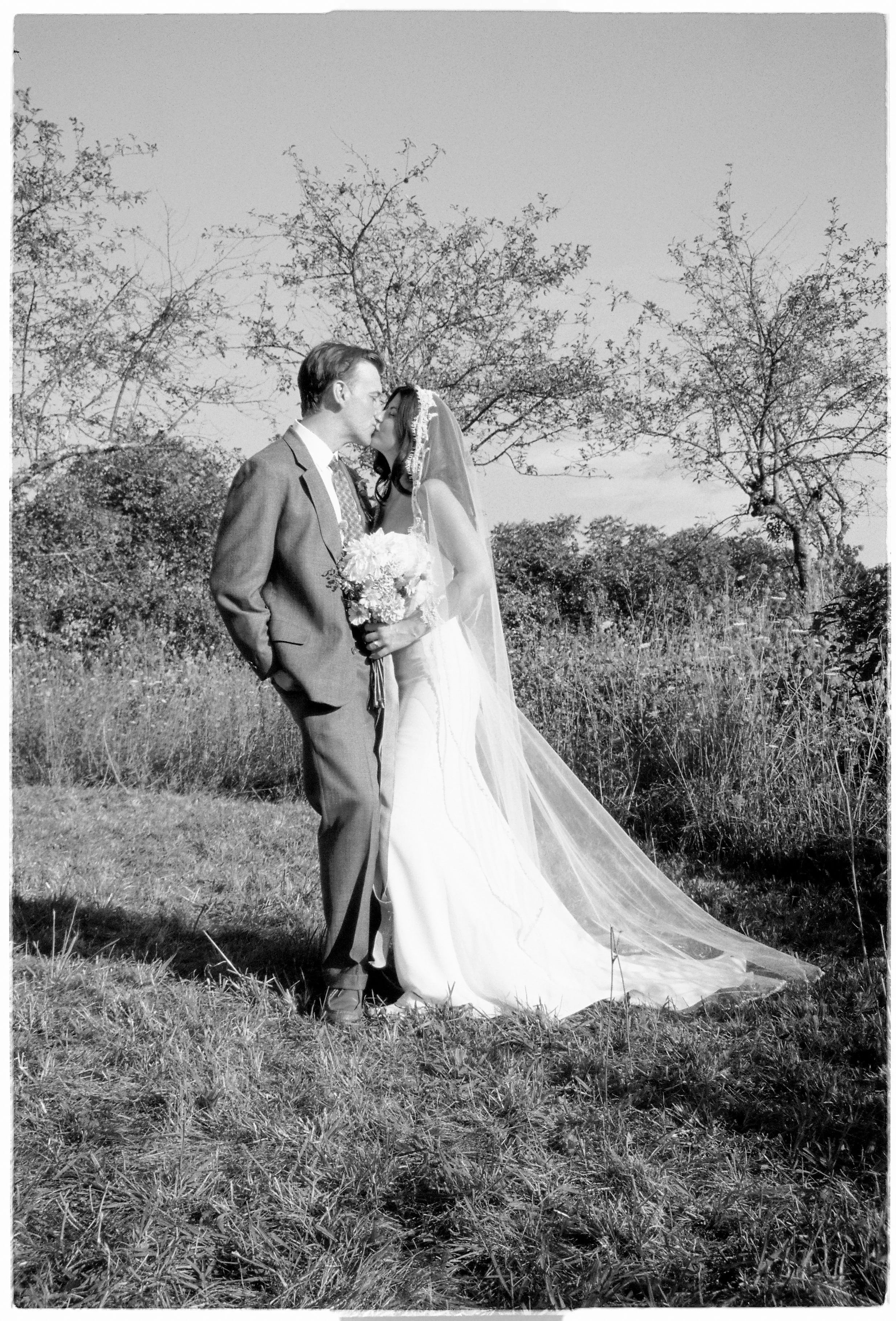 A black-and-white photo of a bride and groom kissing outdoors on a grassy field with trees in the background. The bride wears a wedding dress and veil, holding a bouquet, and the groom wears a suit.