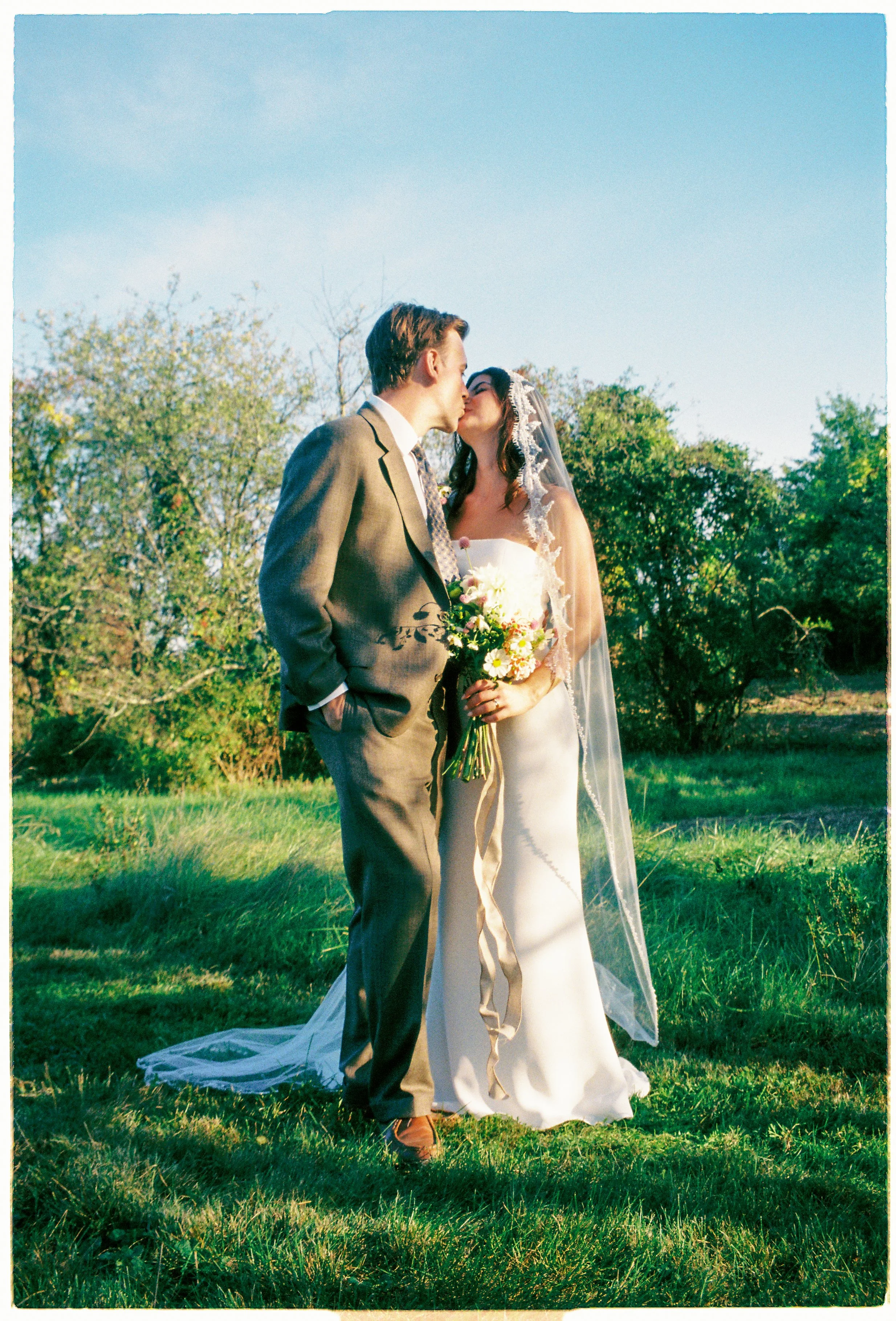 A bride and groom kiss outdoors, standing on grass with trees and a blue sky in the background, during a wedding ceremony.