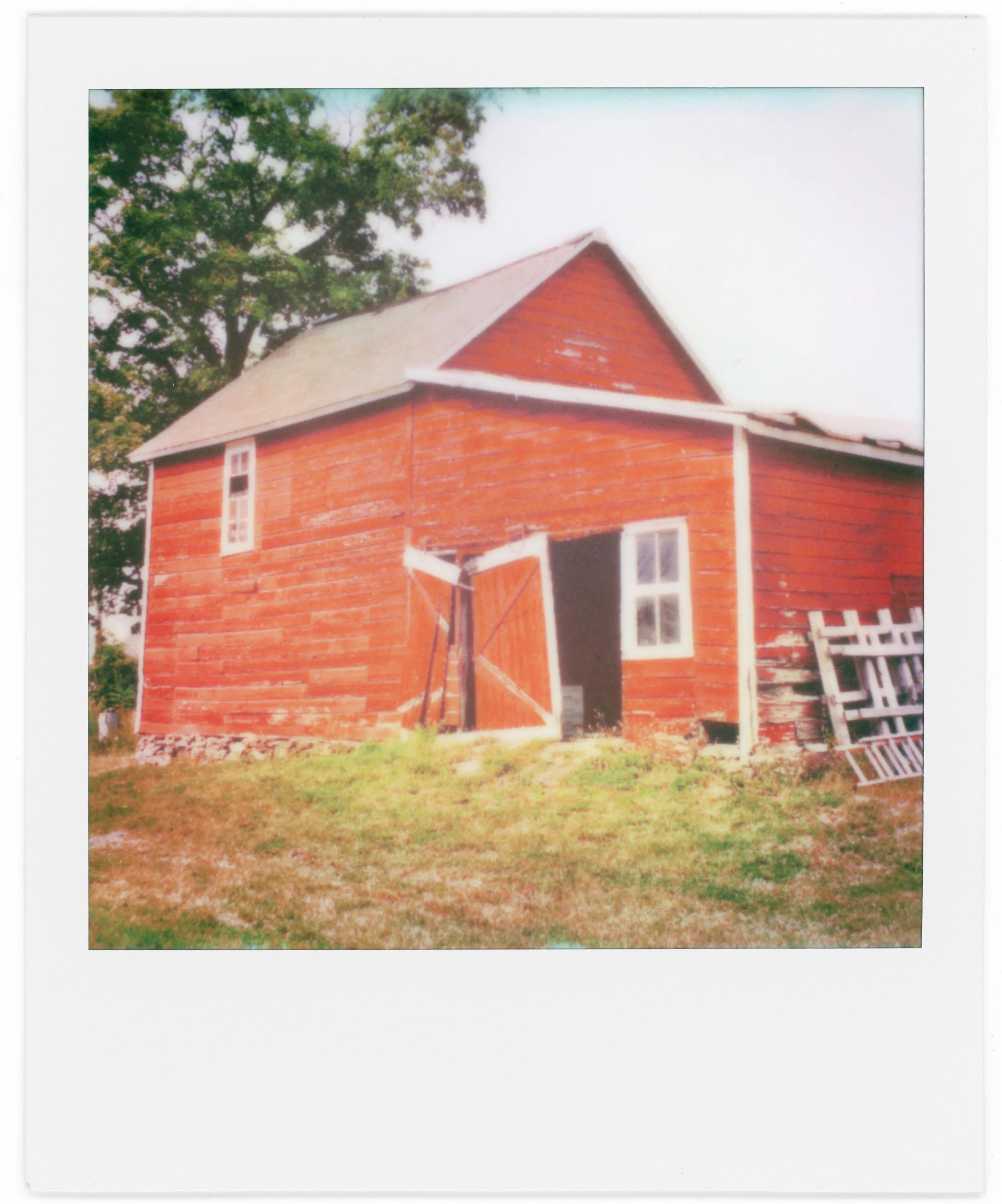 A printed photo of a red barn with white trim, two windows, and a small door, set on a grassy area with trees in the background.