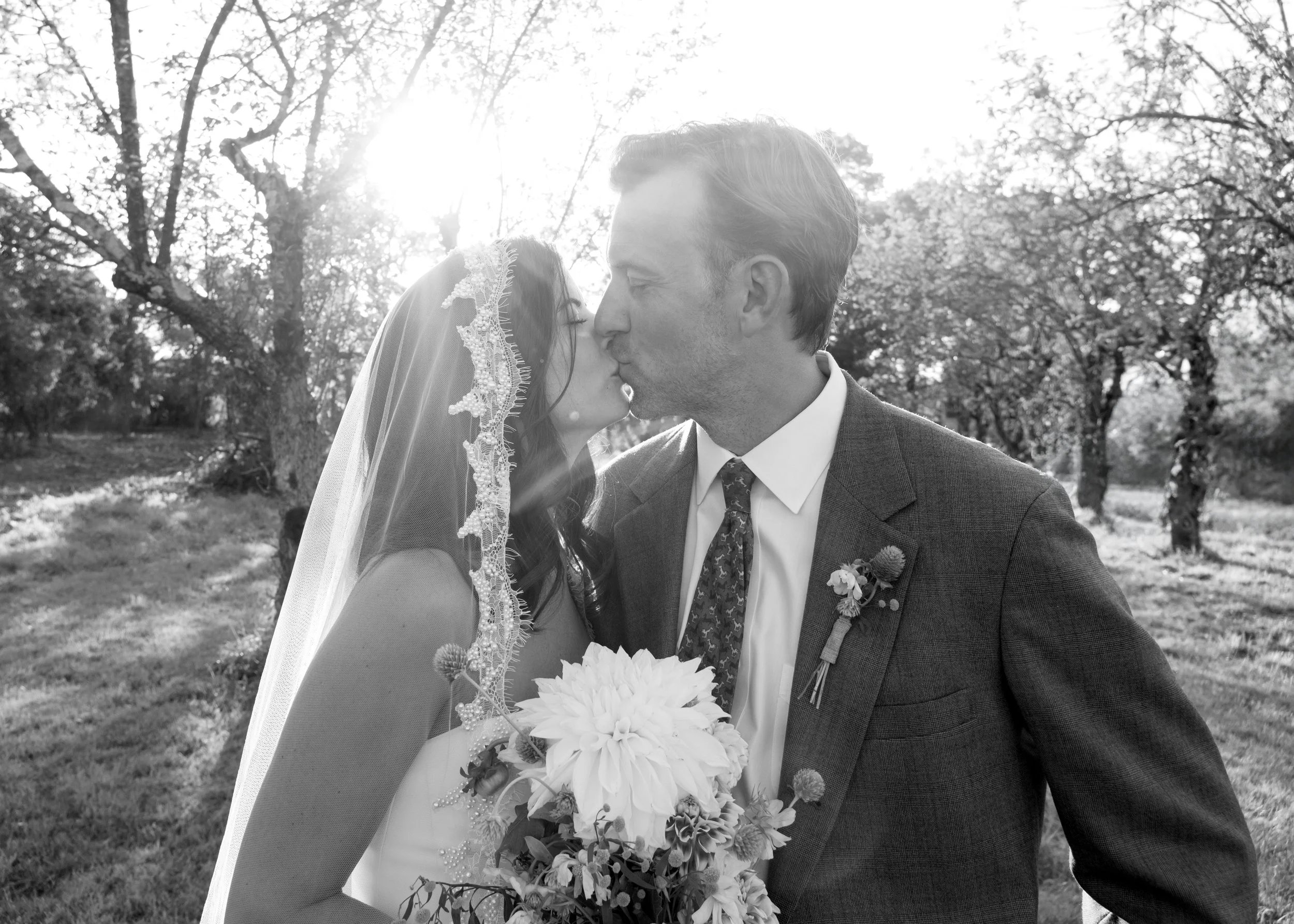 A black and white photo of a bride and groom sharing a kiss outdoors on a sunny day. The bride is wearing a lace wedding veil and holding a bouquet of large flowers, while the groom is dressed in a suit with a patterned tie and boutonnière. The backg