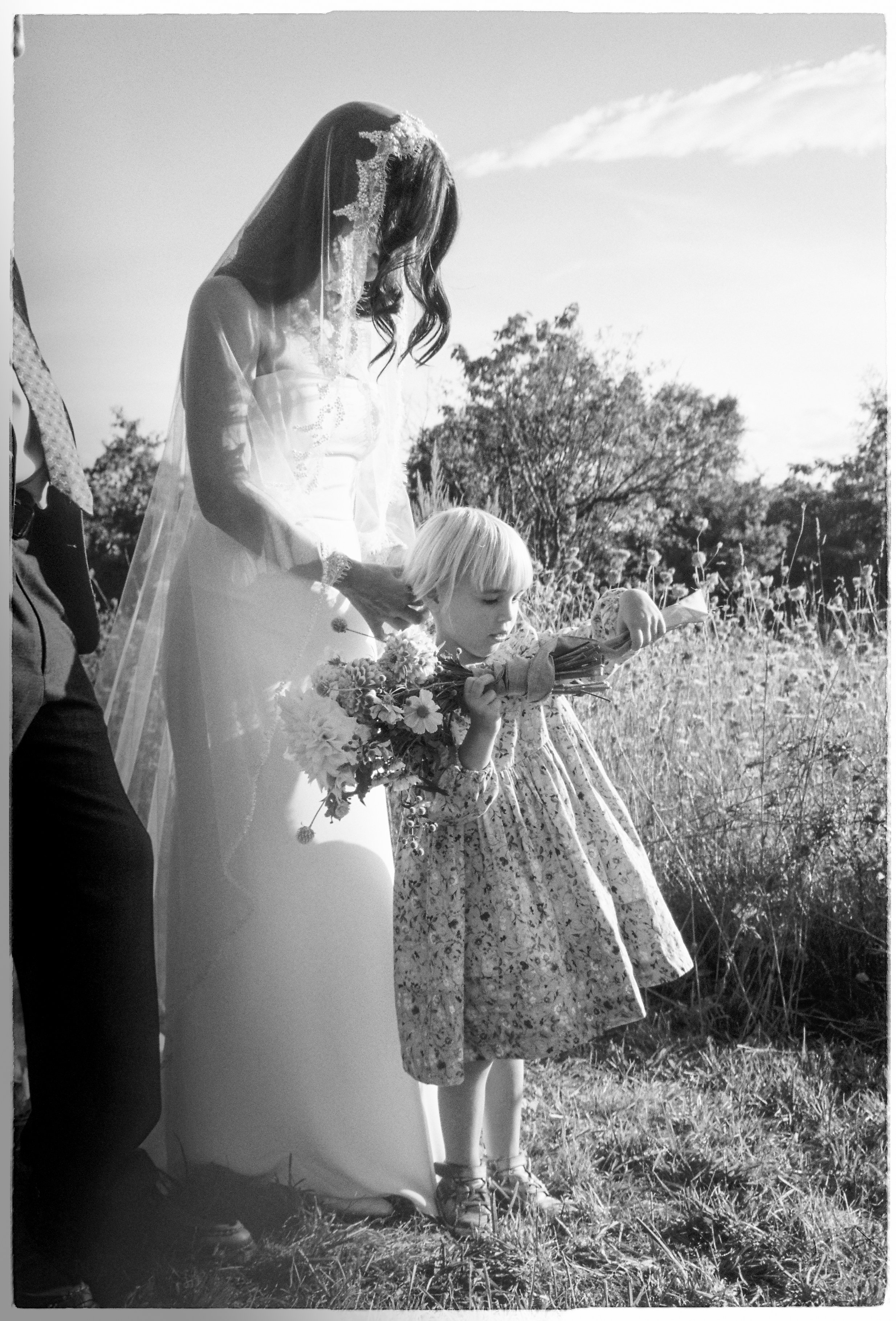 A woman wearing a wedding dress and veil holding a bouquet of flowers, standing outdoors in a field with a young girl in a floral dress pointing at something in the grass.