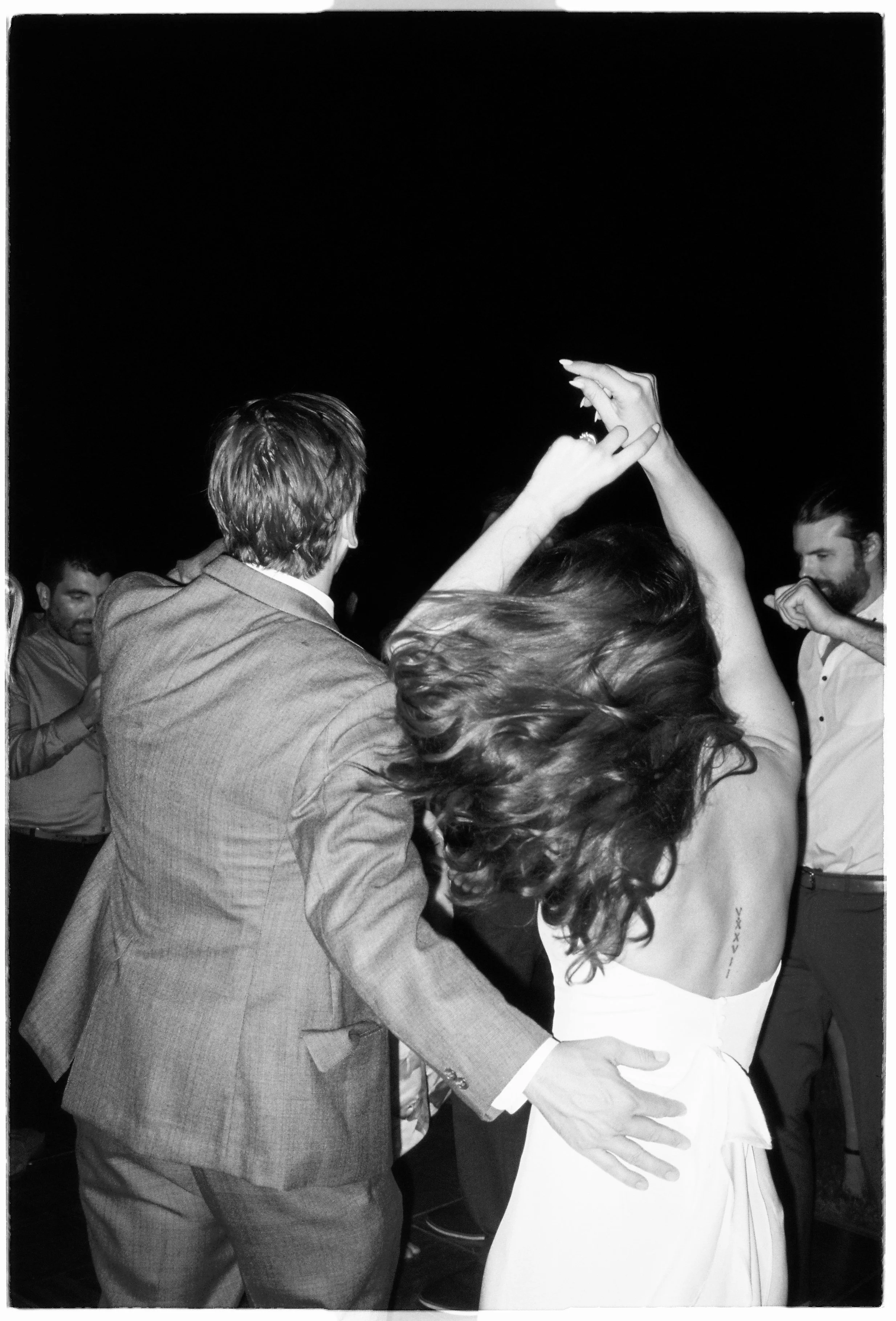 A couple dancing closely at a social event, with the man in a suit and the woman in a strapless dress, surrounded by other people.