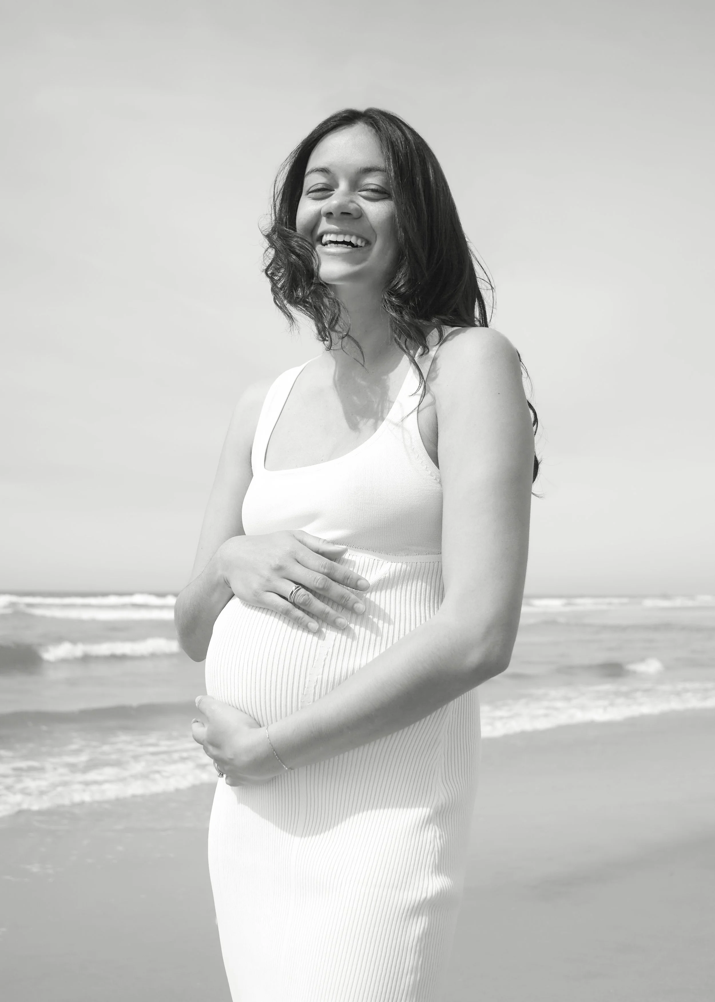 A smiling pregnant woman in a white dress standing on the beach with ocean waves in the background.