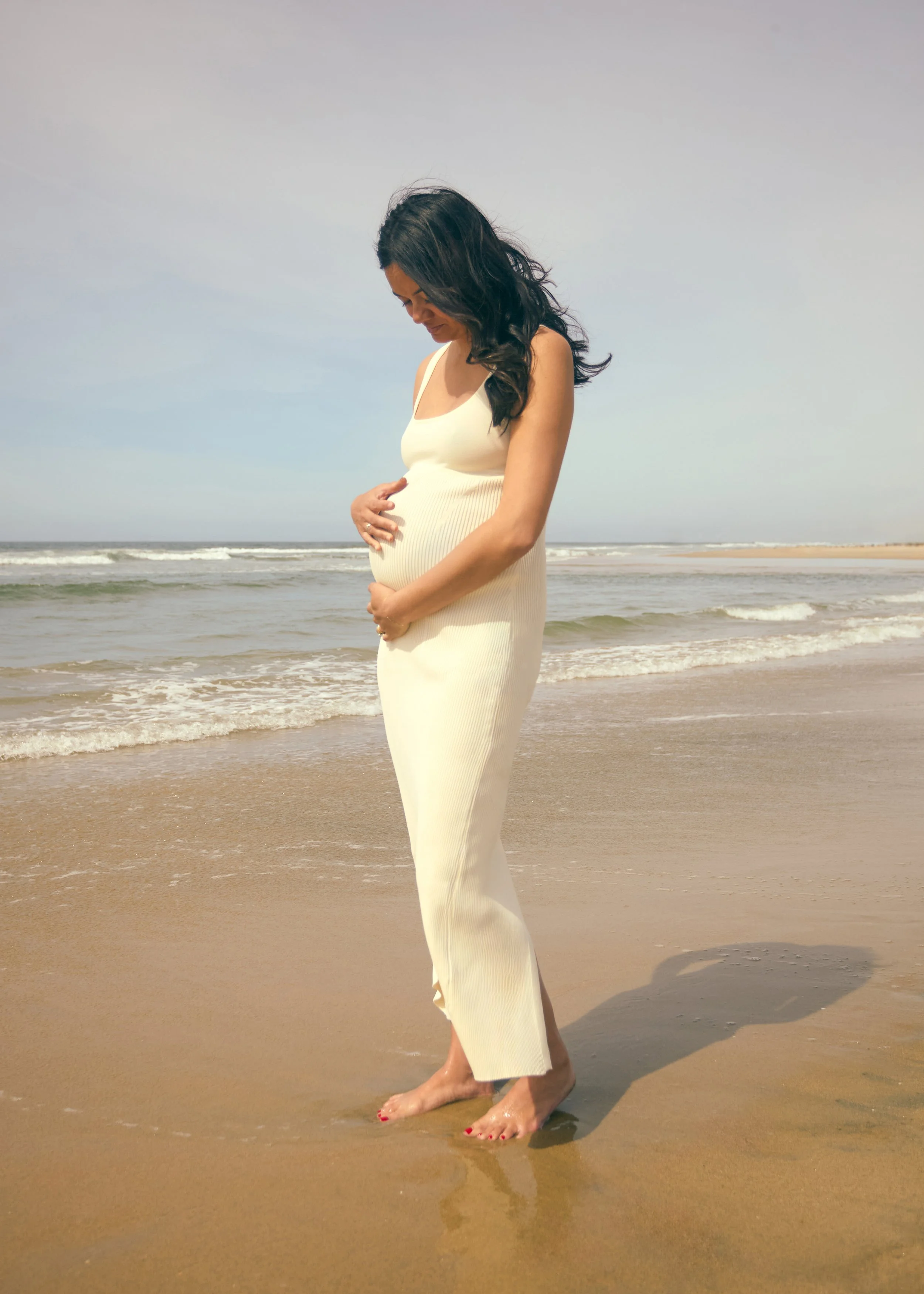 A pregnant woman with long dark hair wearing a white dress standing barefoot on the beach with small waves in the background.
