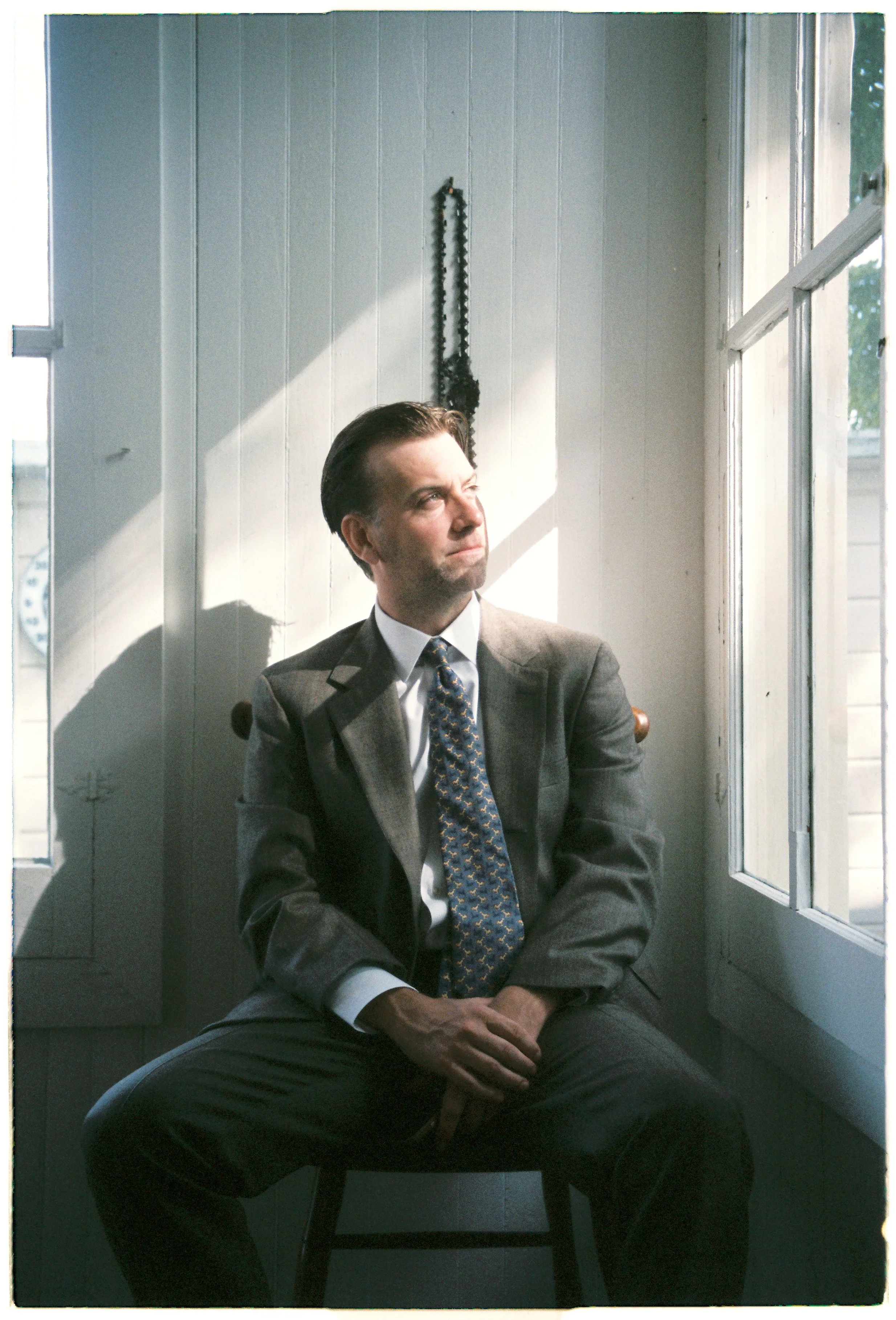 A man in a gray suit and patterned tie sitting in a room with white paneled walls and an open window, looking thoughtfully to the side.