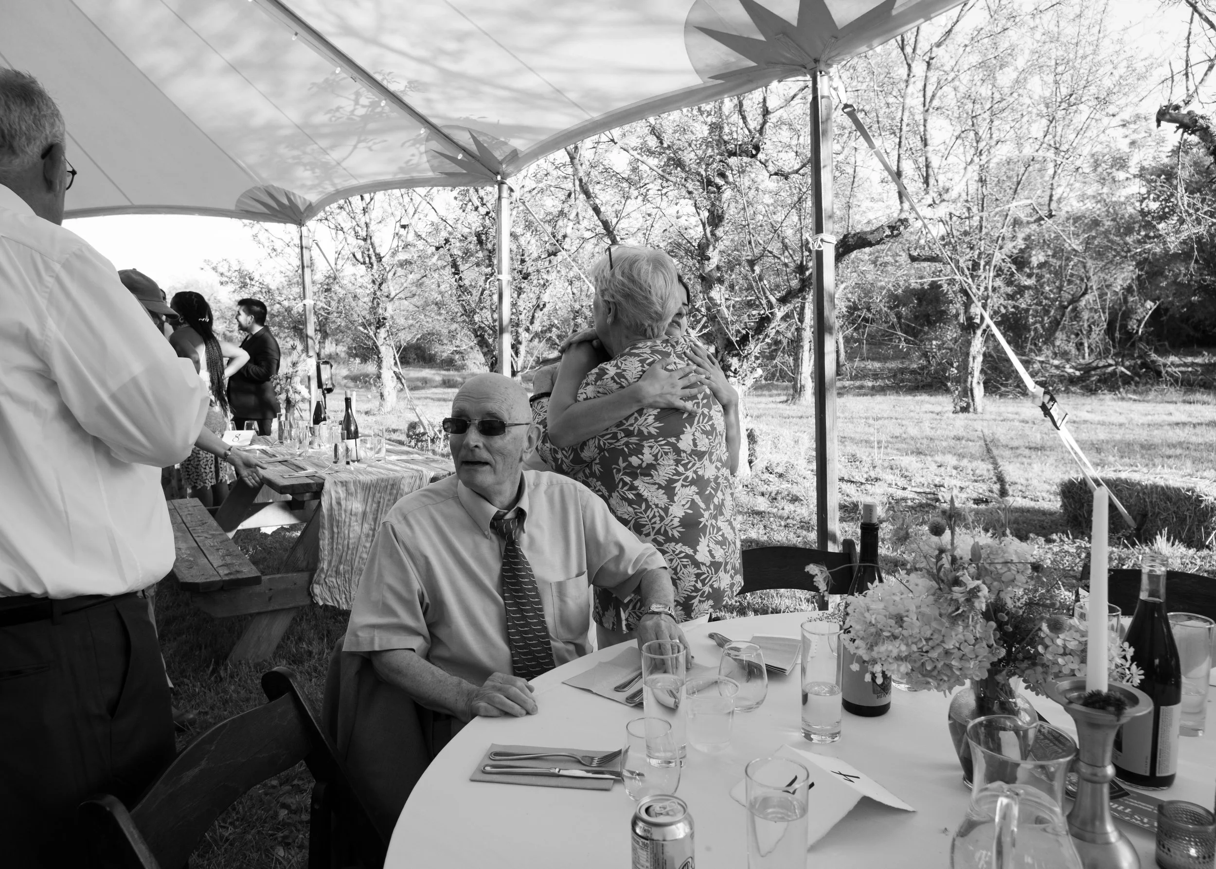 People attending a gathering outdoors under a canopy, with tables set for a meal, including wine bottles, glasses, and a floral centerpiece.