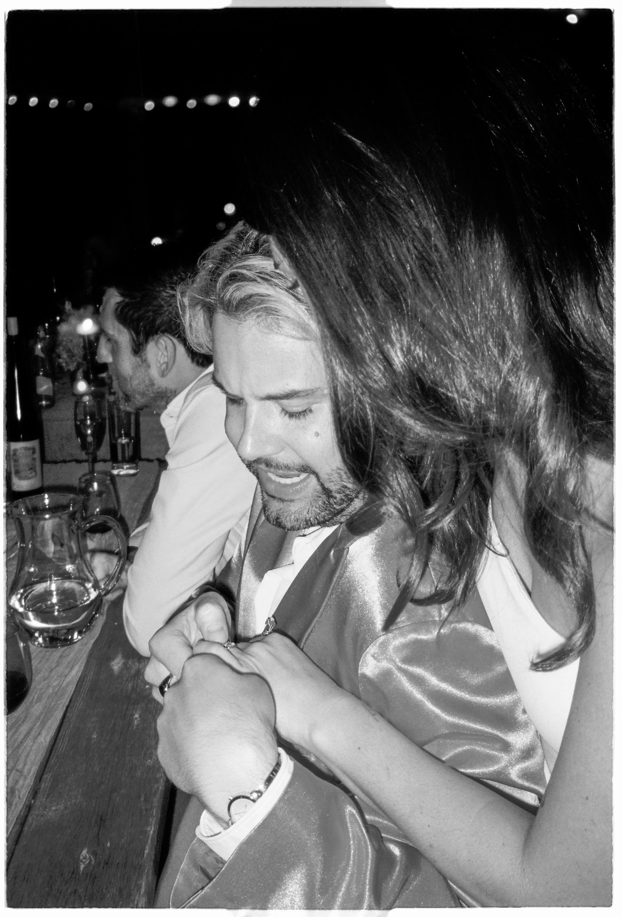A group of people, including a man with long hair and a woman with a bracelet, sitting at a bar or restaurant table, engaged in conversation, with drinks and bottles on the table.