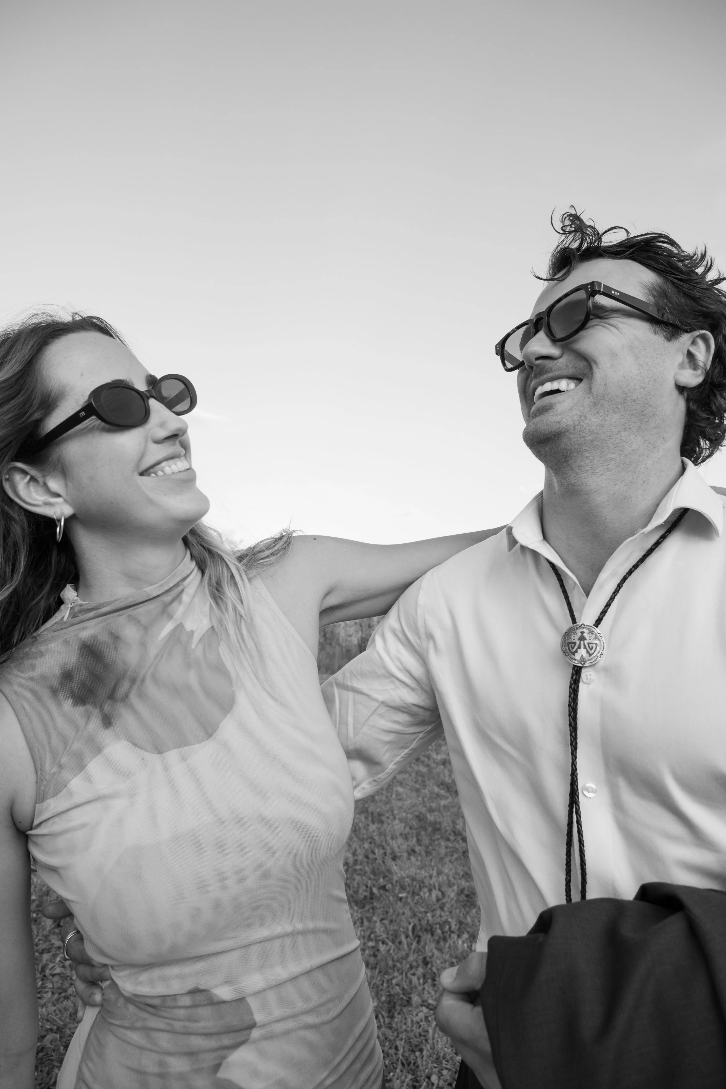 A black and white photo of a smiling man and woman outdoors, both wearing sunglasses, with their arms around each other.