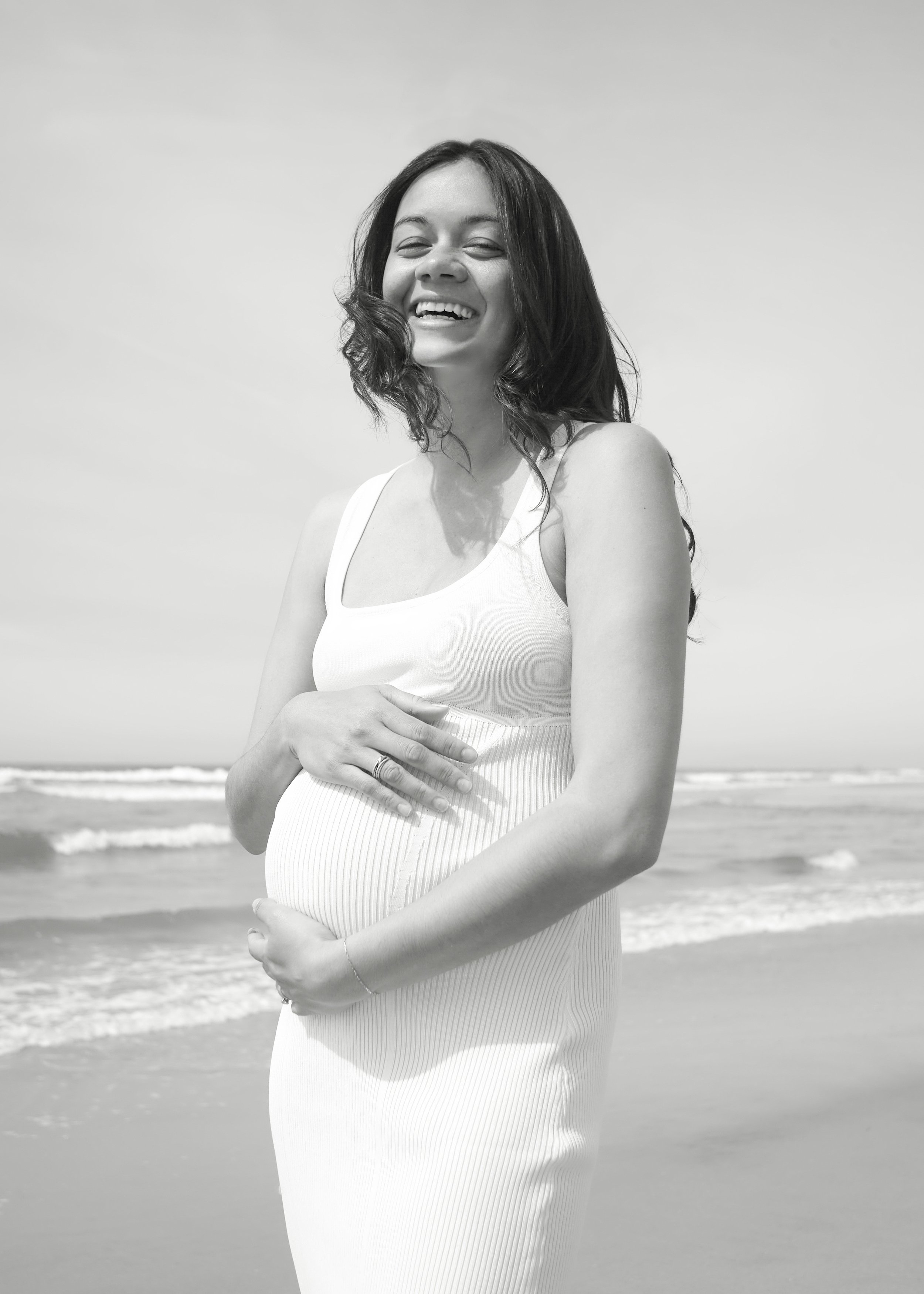 Black and white photo of a pregnant woman smiling at the beach, holding her belly with both hands, wearing a sleeveless dress.