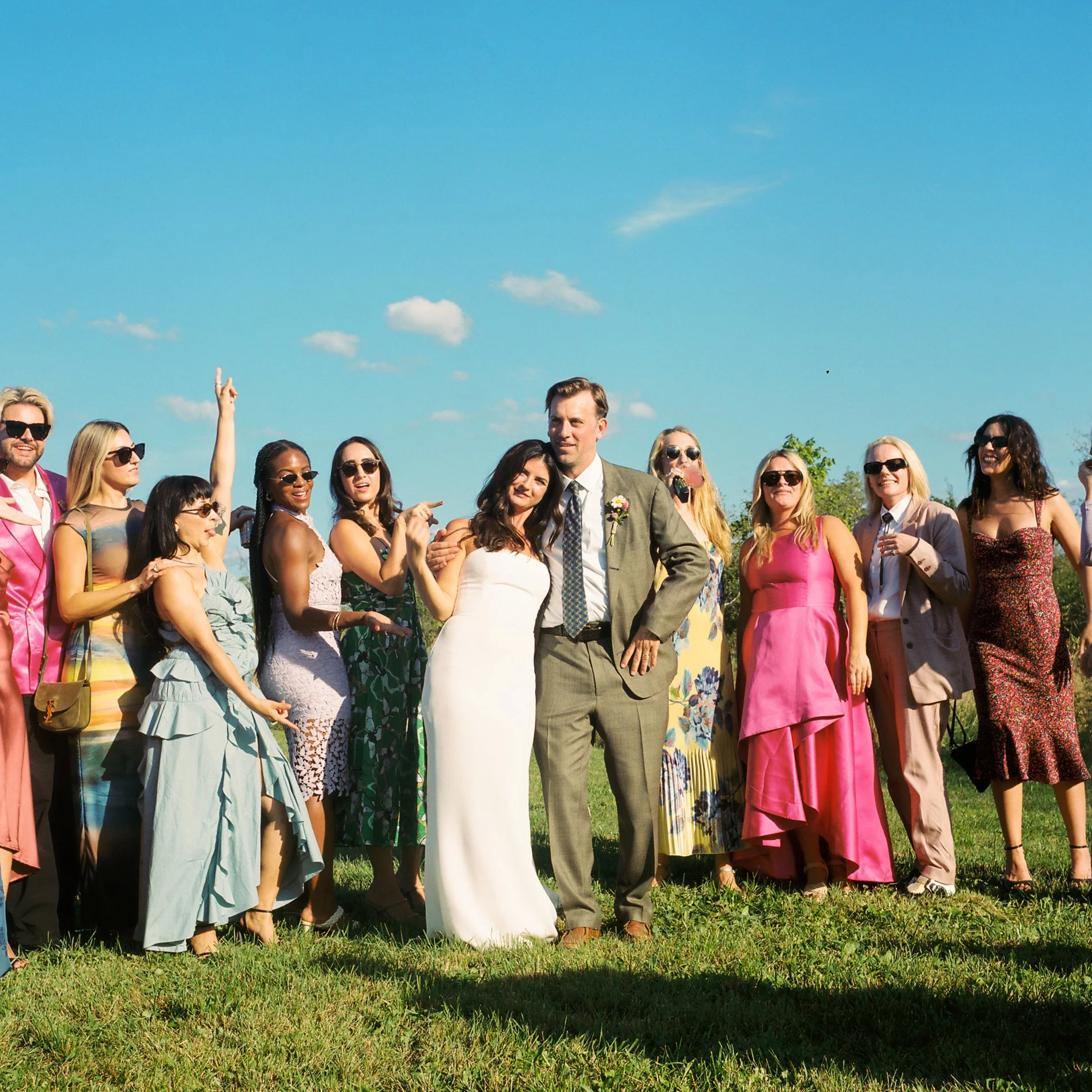 Group of diverse people dressed in colorful clothing, standing outdoors on grass under a blue sky with some clouds, celebrating or socializing.
