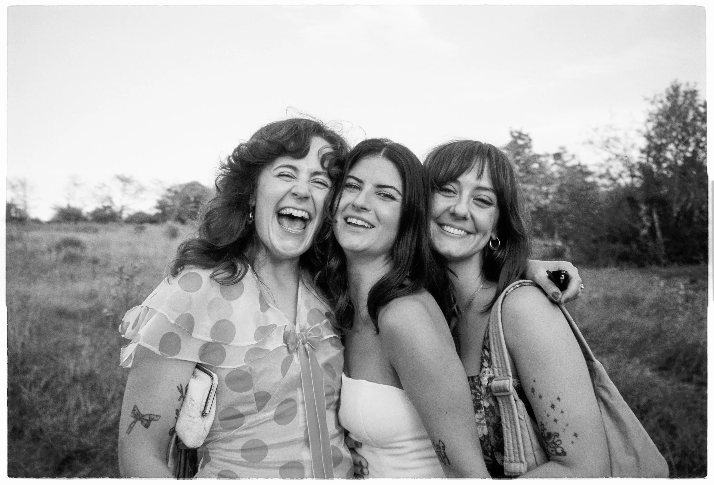 Three women standing outdoors in a field or park, smiling and enjoying each other's company. One woman on the left is laughing with her mouth open, the middle woman has straight dark hair and a light-colored top, and the woman on the right has dark h