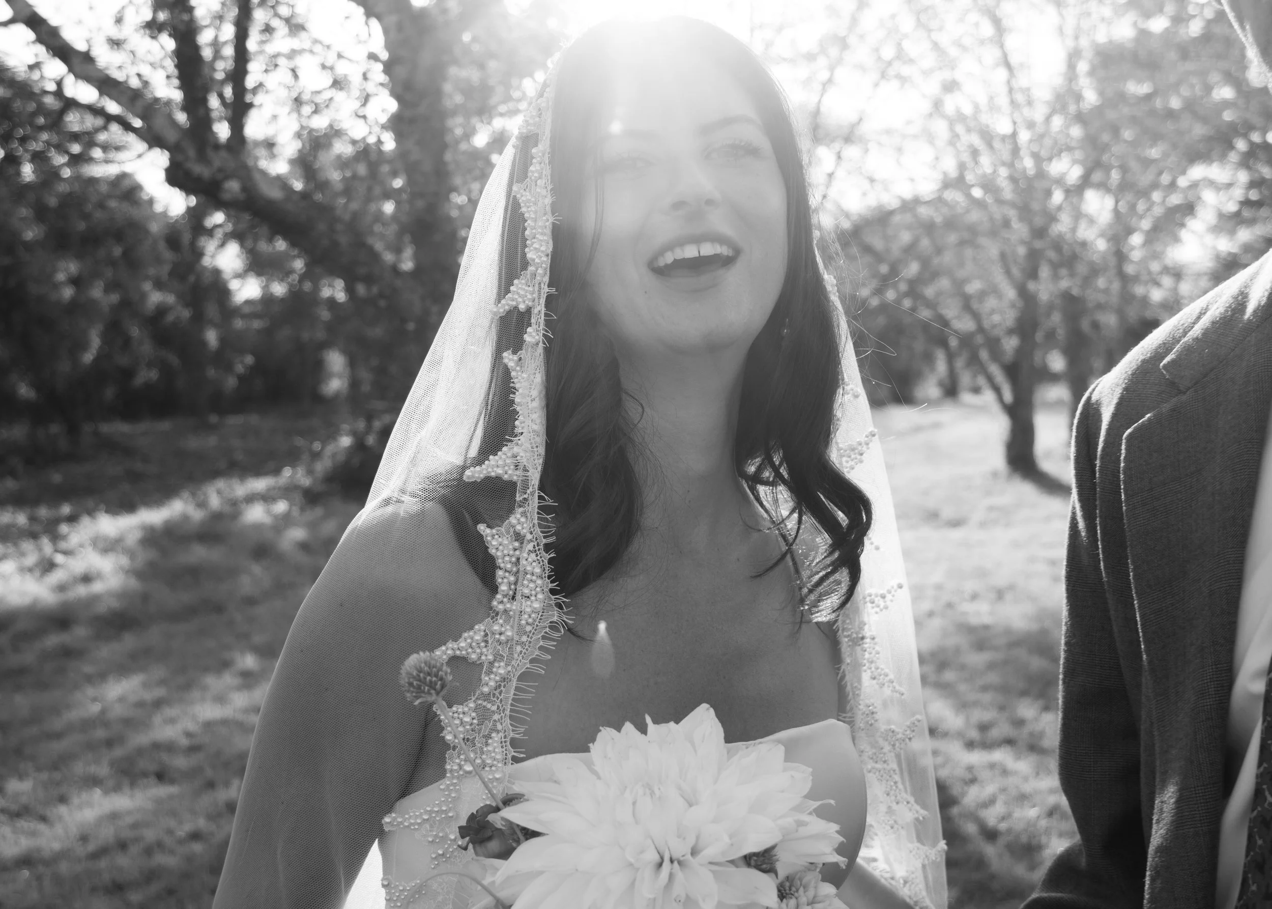 Black and white photo of a woman wearing a wedding dress and veil, holding a bouquet of flowers, outdoors with trees in the background, smiling with her mouth open.