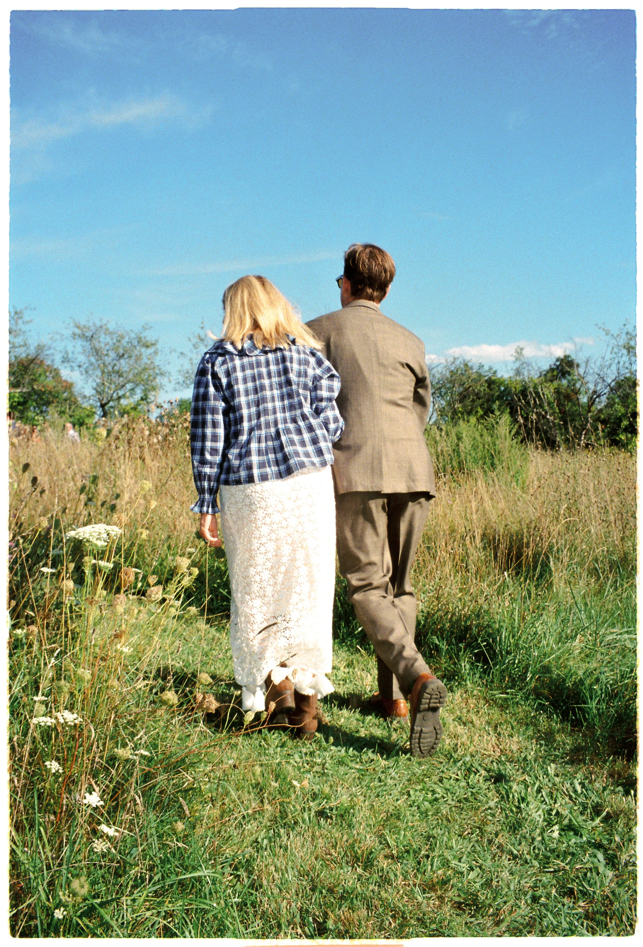 A woman with blonde hair wearing a blue checkered shirt and a long white skirt, and a man in a brown suit and glasses, walk together through a grassy field under a clear blue sky.