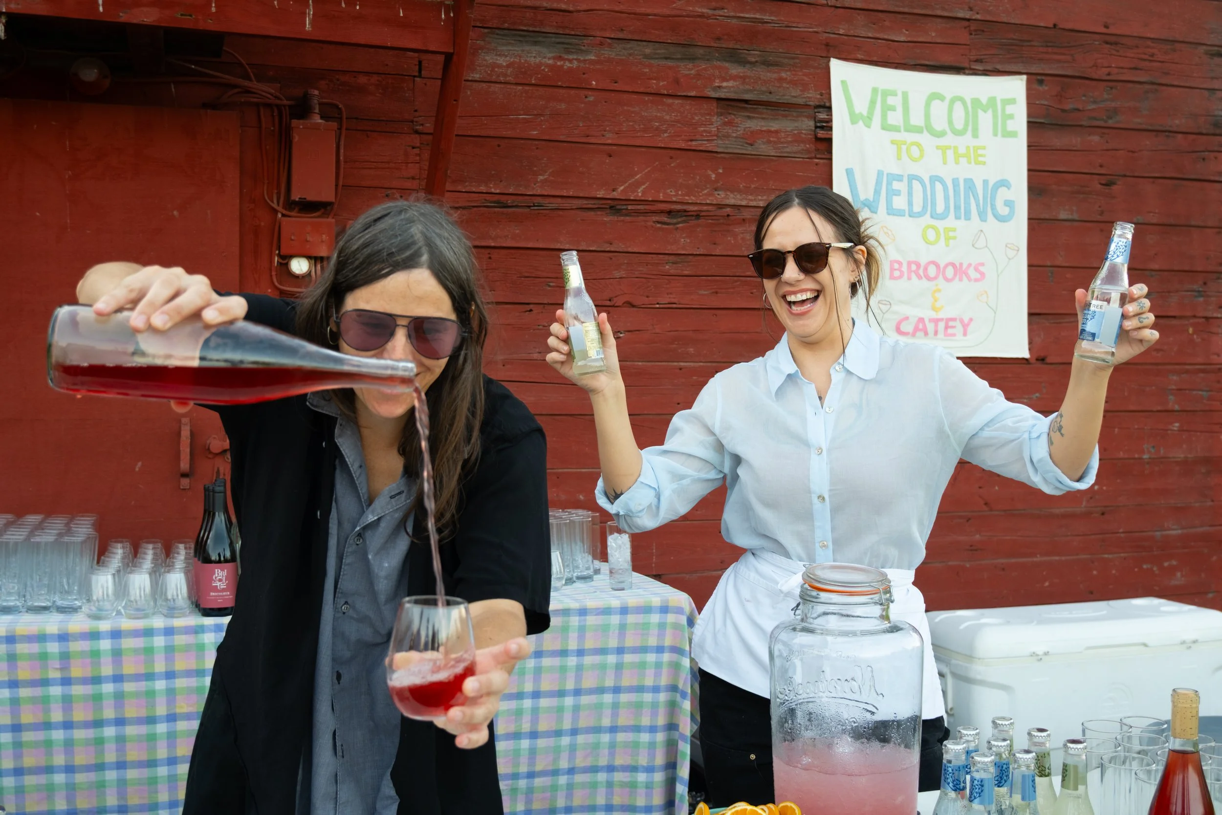 Two women at an outdoor wedding reception, one pouring red liquid into a glass, the other holding a bottle and smiling, with a colorful sign in the background that reads "Welcome to the Wedding of Brooks & Catey".