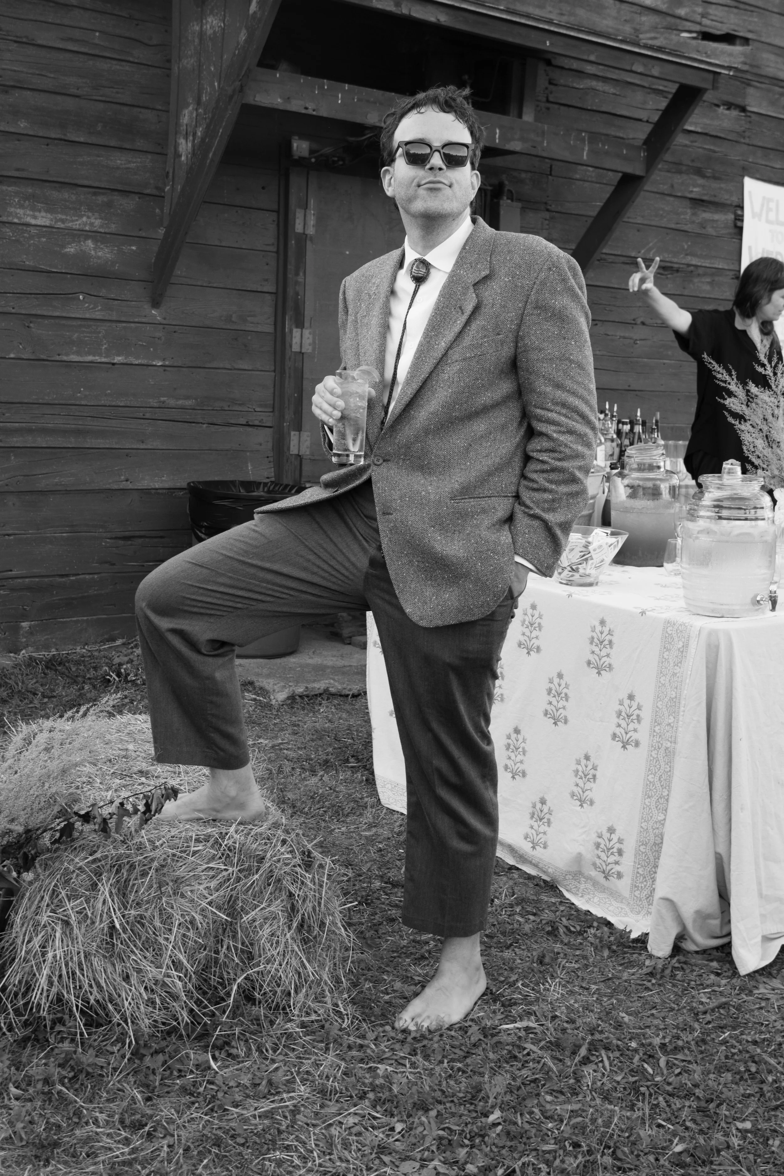 A man in a suit with sunglasses holding a drink, standing barefoot on grass next to a hay bale at an outdoor event with a wooden building in the background. A woman behind a table with jars and bottles is gesturing with peace signs.