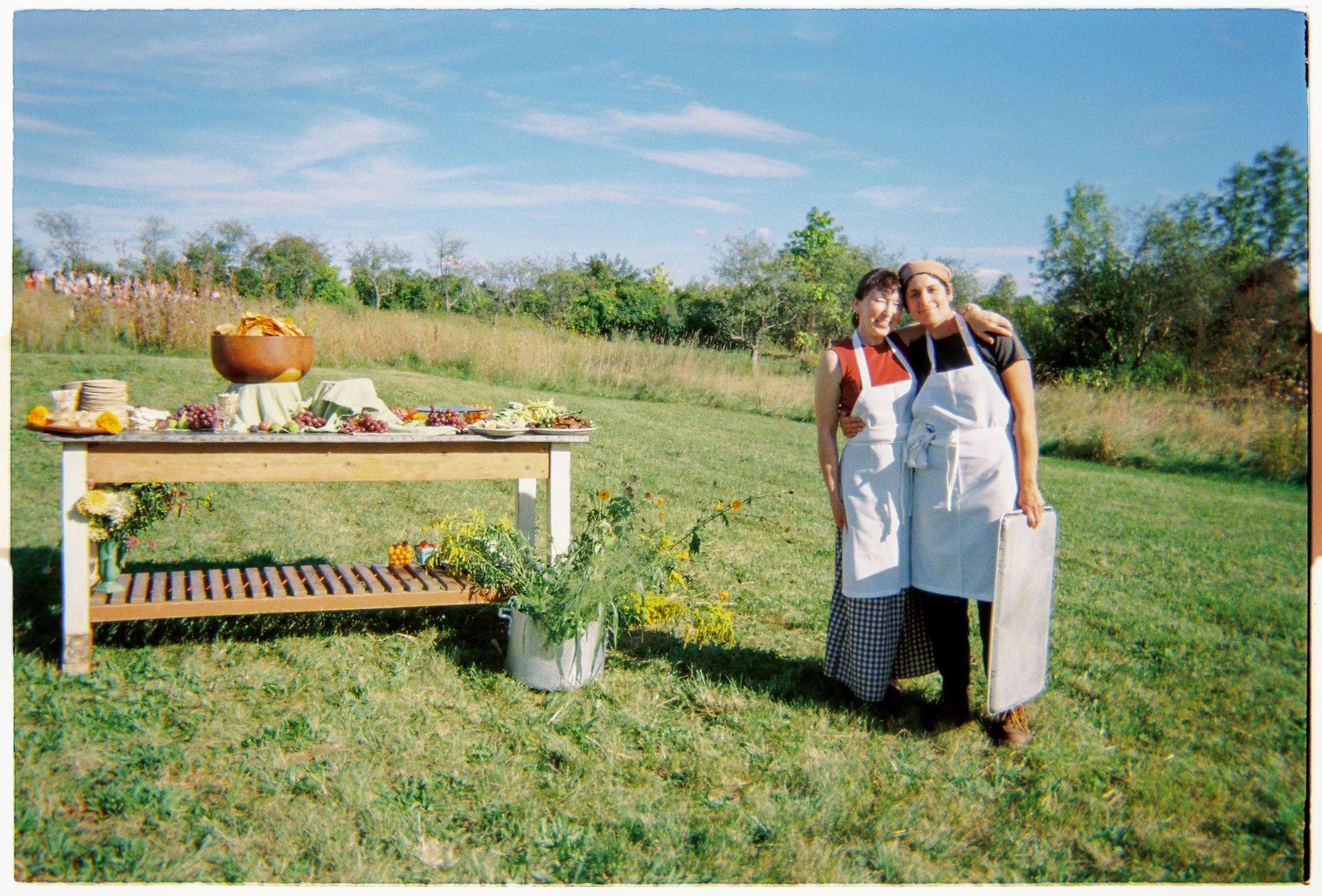 Two women wearing aprons standing outdoors near a table with food, in a grassy area with trees and blue sky in the background.