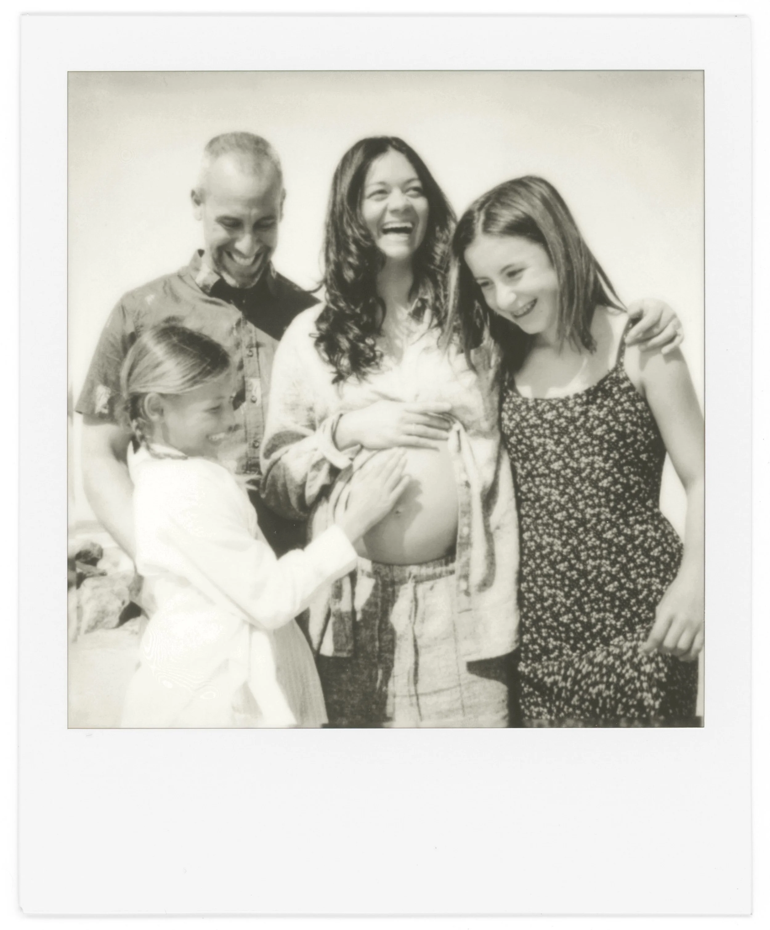 A black-and-white photograph of five people smiling and sharing a joyful moment, standing close together, with a plain background, captured with a Polaroid camera.