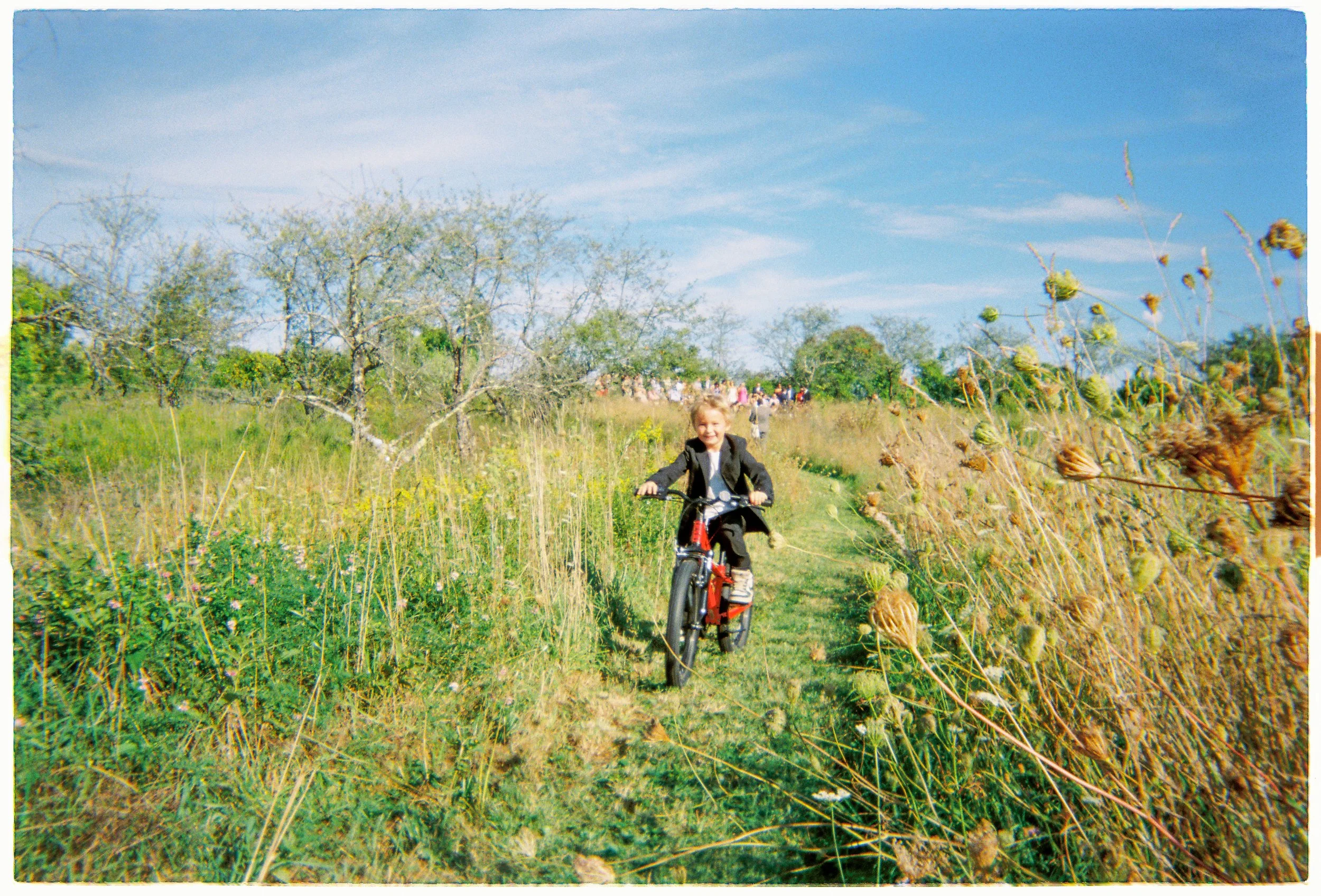A young child riding a bicycle along a grassy trail surrounded by wildflowers and trees on a bright, sunny day with a blue sky.