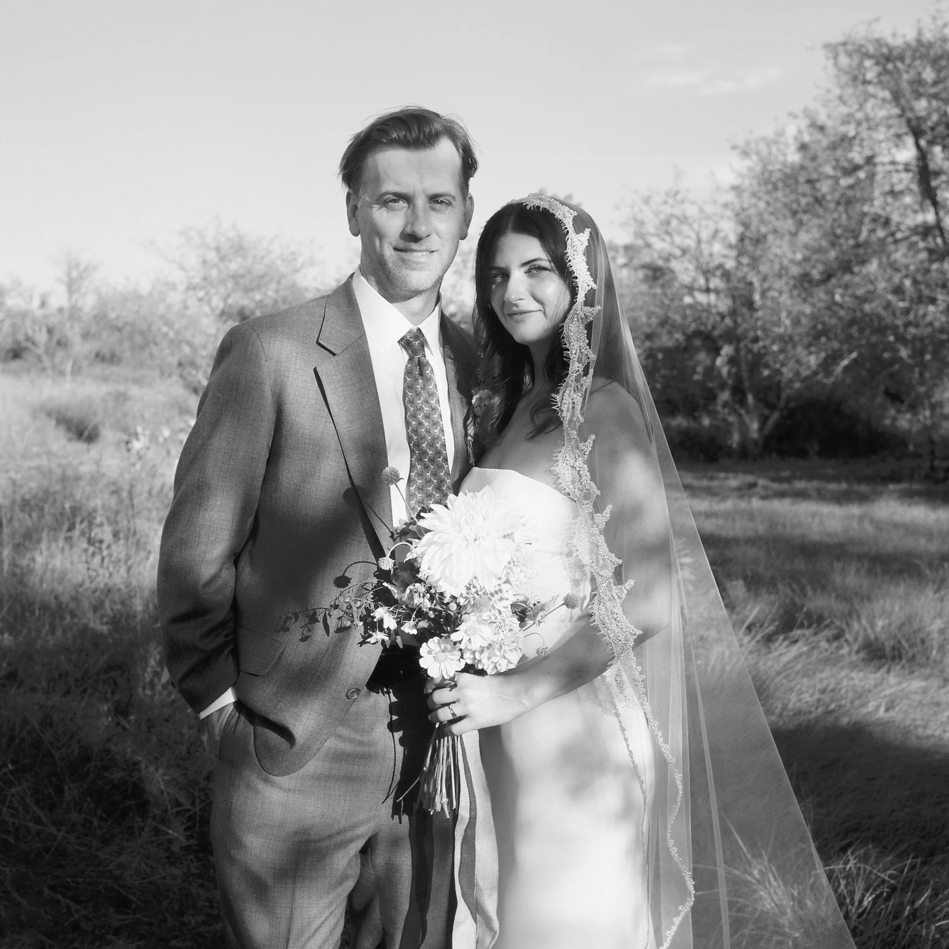 Black and white photograph of a newlywed couple outdoors, smiling at the camera. The groom wears a suit and tie, and the bride is in a wedding dress with a lace veil, holding a bouquet of flowers. Background includes trees and open sky.