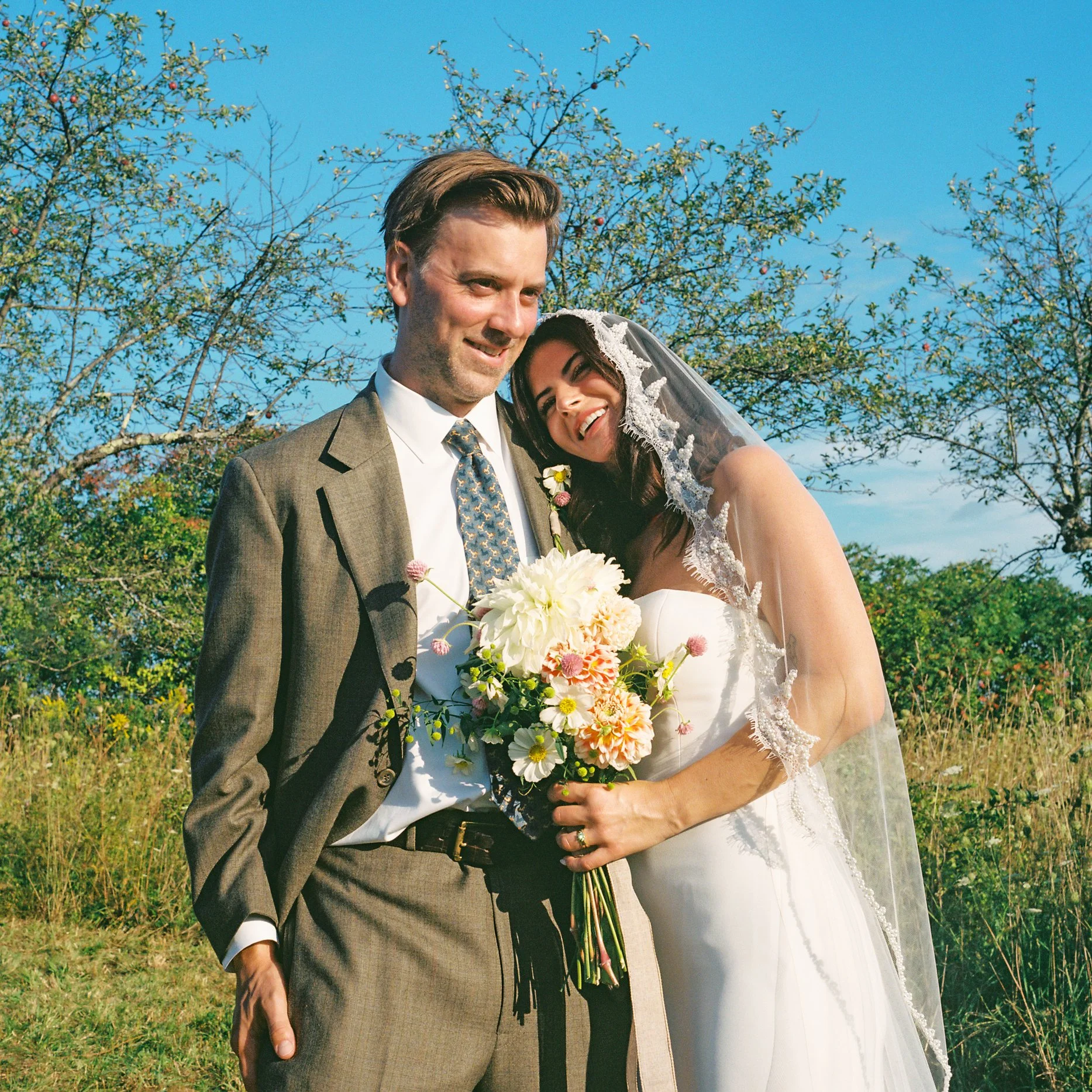 A smiling bride and groom posing outdoors on their wedding day, with trees and blue sky in the background. The bride is holding a bouquet of white and pink flowers and leaning her head on the groom's shoulder.