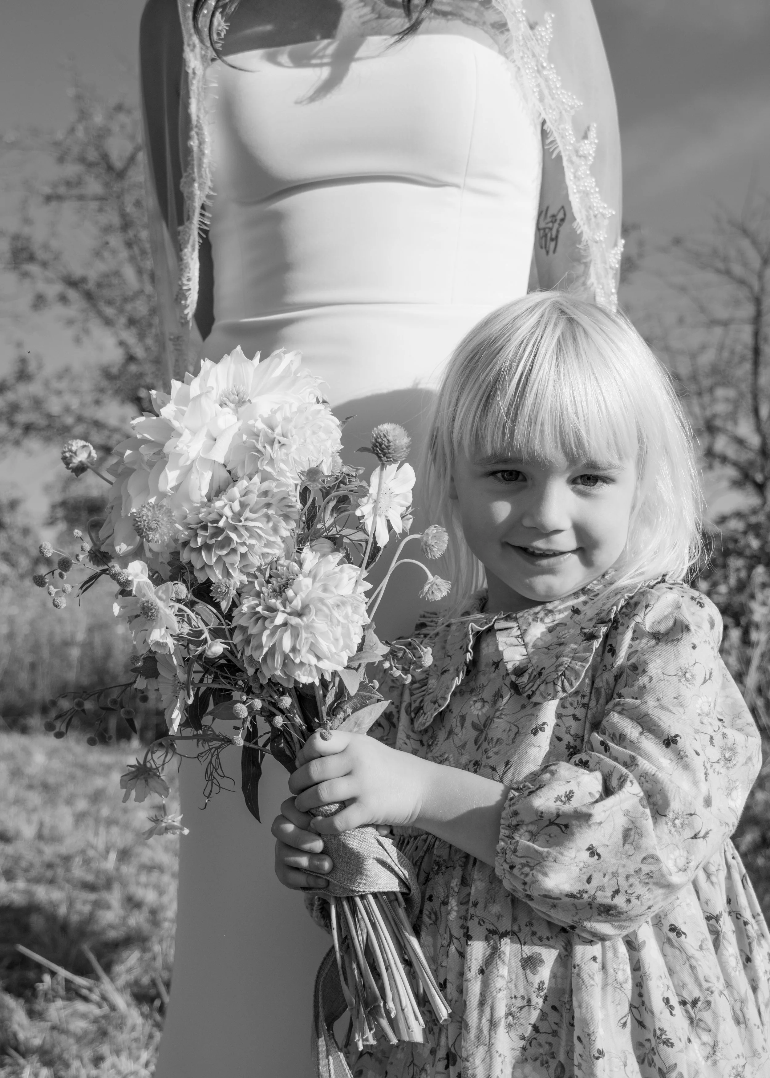 A young girl with straight blonde hair holding a bouquet of flowers, standing outdoors, with a woman in a wedding dress in the background.