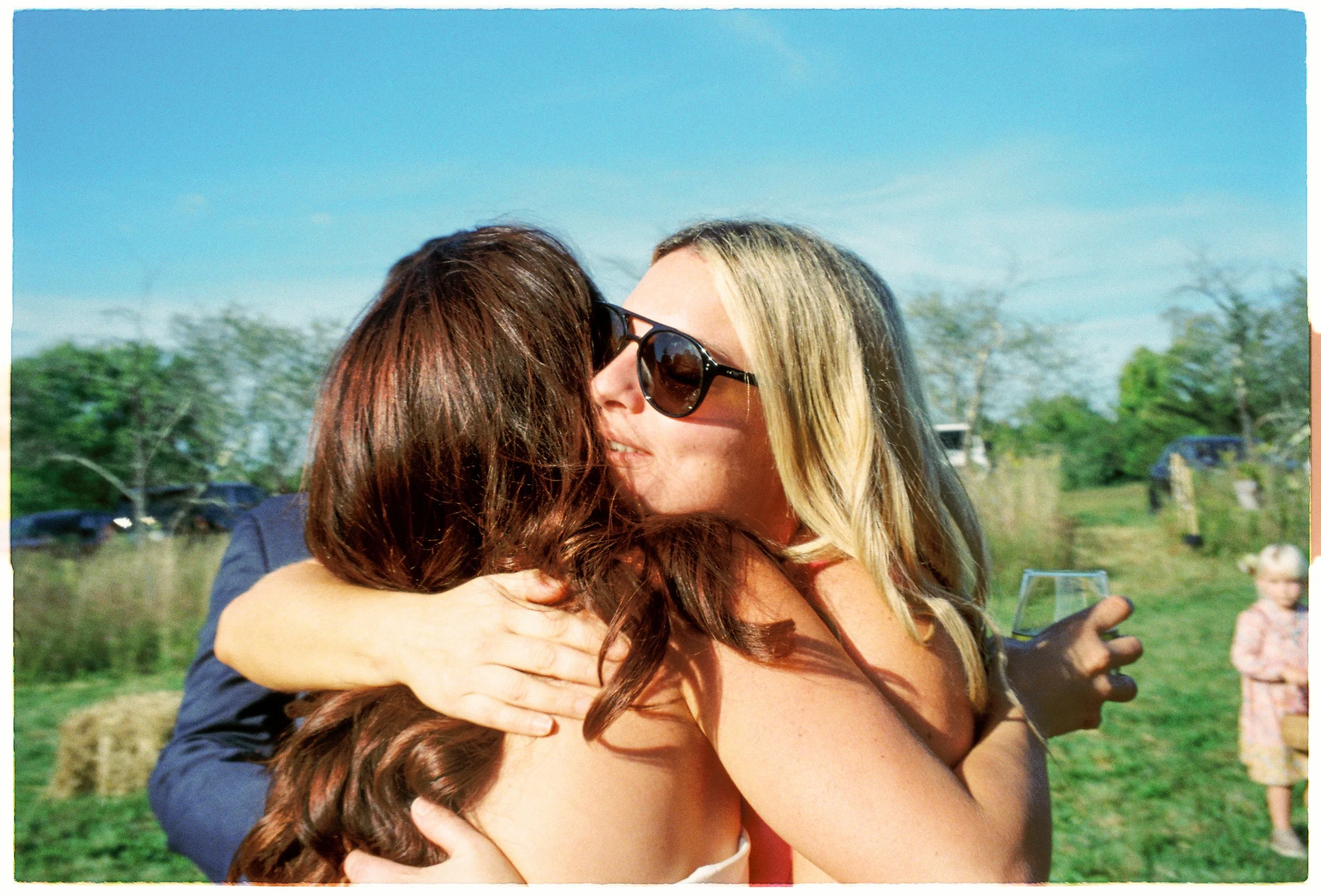 Two women hugging outdoors on a sunny day, one wearing sunglasses and the other with long brown hair, with people and trees in the background.