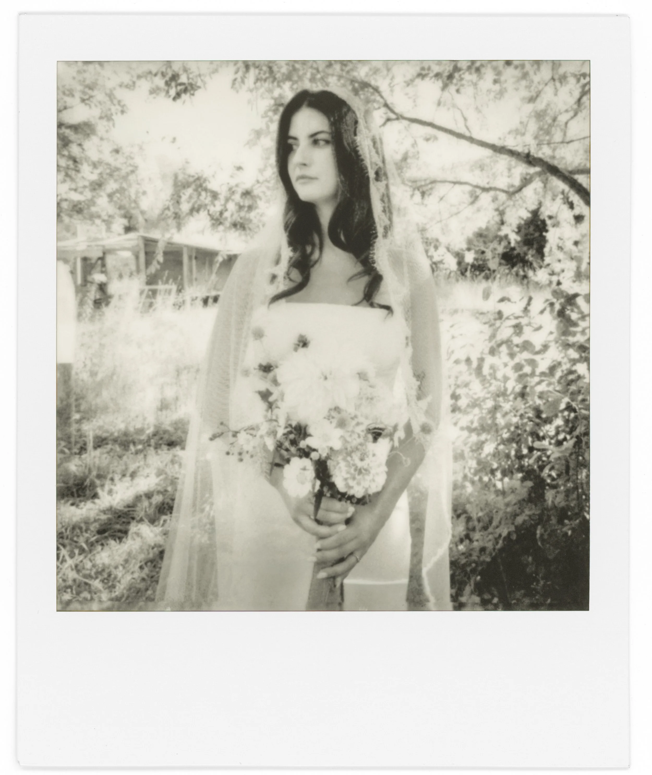 Black and white photograph of a woman with long dark hair holding a flower bouquet, standing outdoors in a natural setting with trees and foliage in the background, framed with a white border.