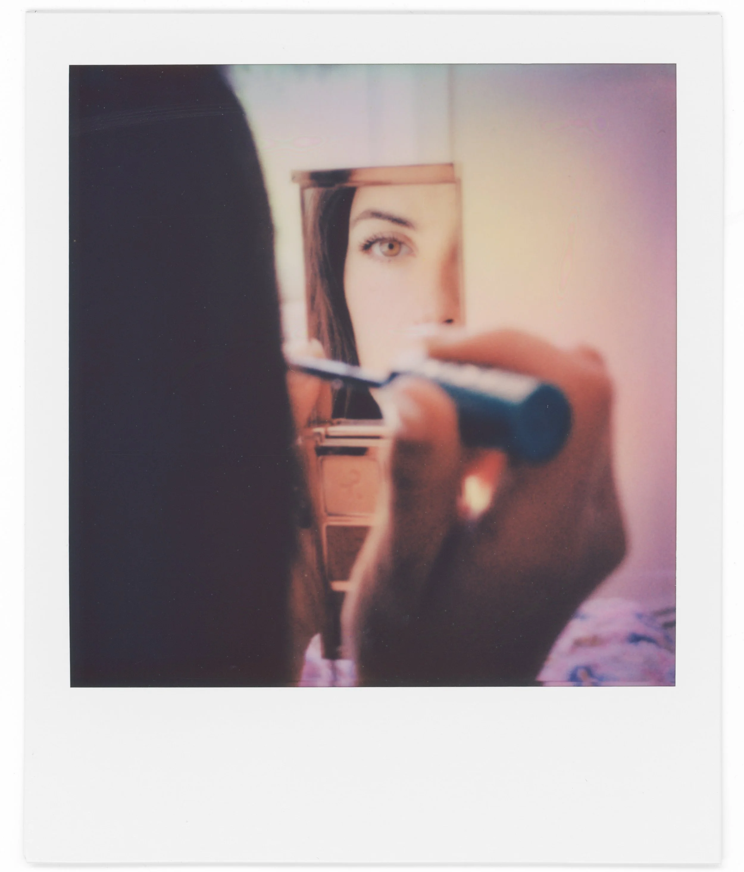 A Polaroid-style photo showing a woman with brown hair and light-colored eyes looking into a mirror and taking a selfie. The background has a gradient with pink and purple hues.