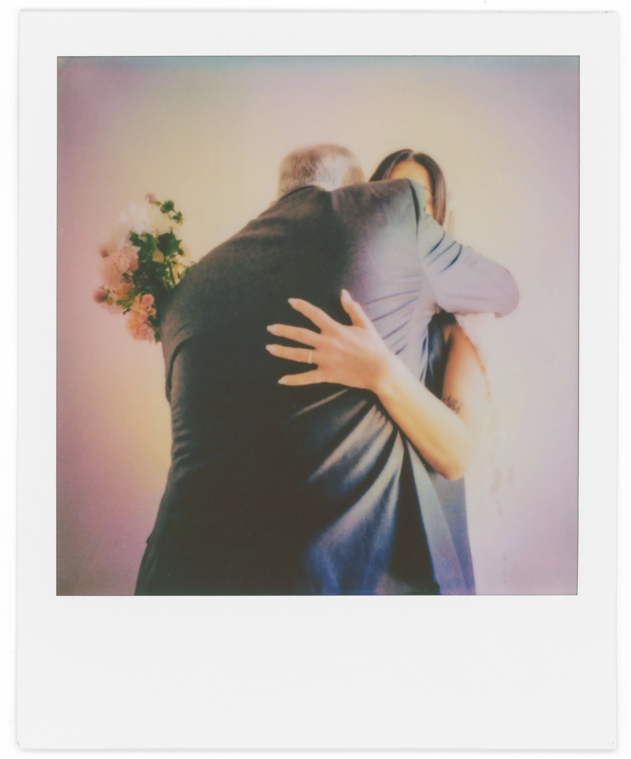 A Polaroid photo of a man and woman hugging each other closely, with the woman's hand visible on the man's back, and behind them a vase with white and pink flowers. The background is a plain, light-colored wall.