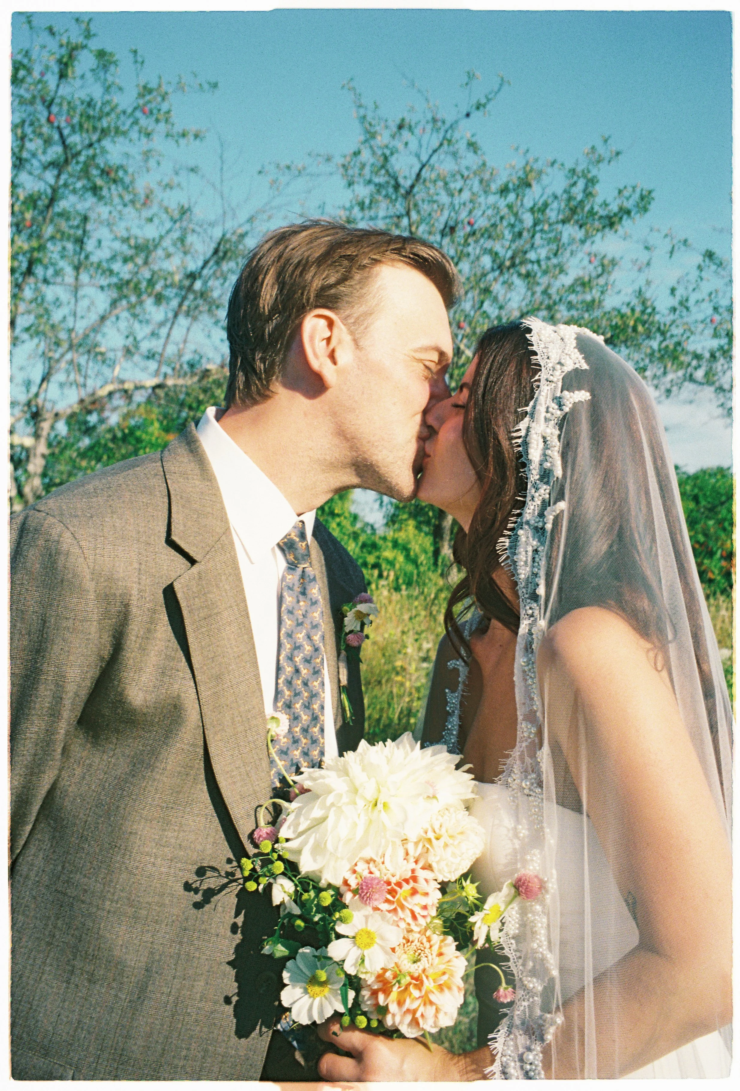 A couple, dressed in wedding attire, kissing outdoors on a sunny day with trees and blue sky in the background. The bride is holding a colorful bouquet.