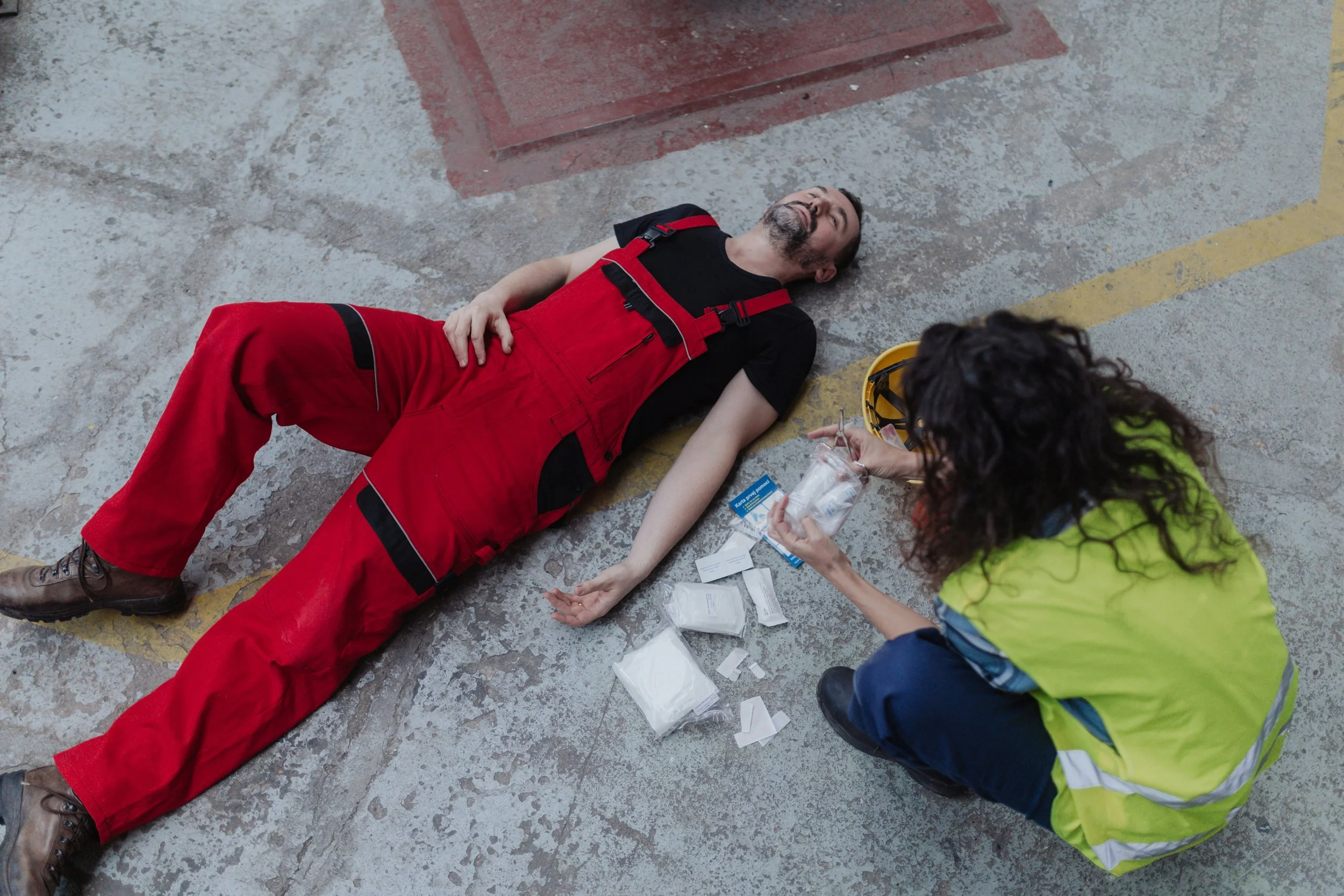 A man lying on the ground receiving medical attention from a woman. The man is wearing red work overalls and a black t-shirt, and appears to be injured or unconscious. The woman, wearing a yellow safety vest, is holding medical supplies and administering aid. The scene appears to be in a parking lot or industrial area with concrete ground.