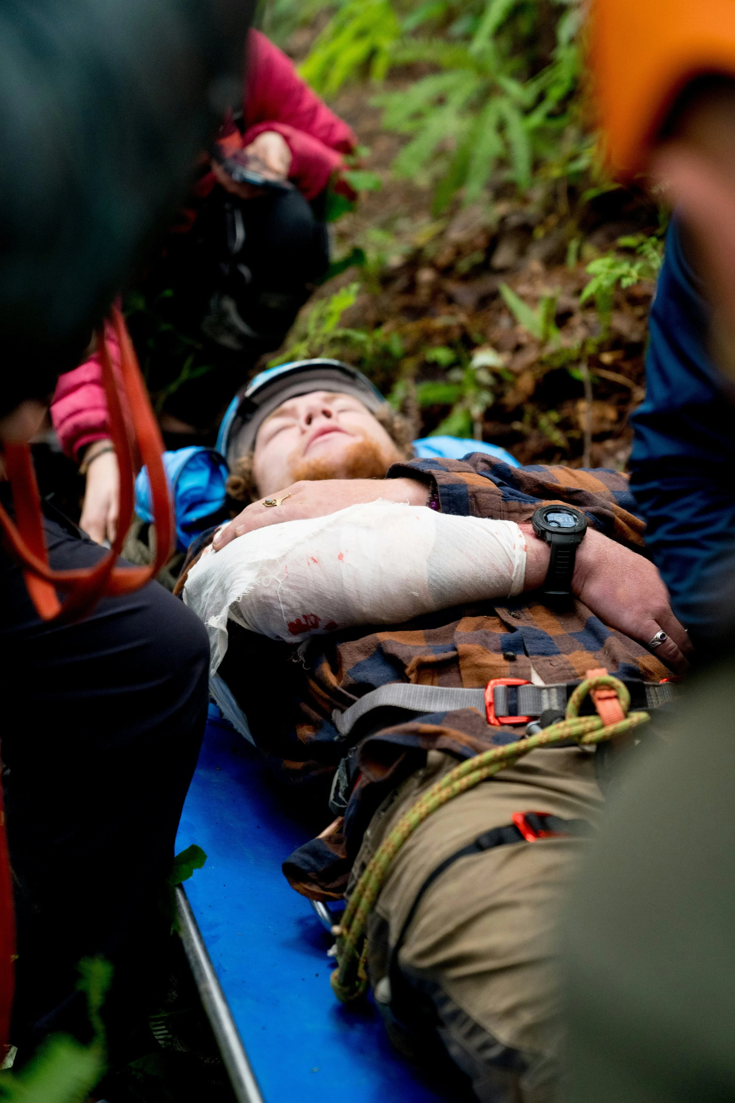 A person with a beard and curly hair lying on a blue rescue stretcher in a forested area, wearing a helmet and harness, with others assisting.