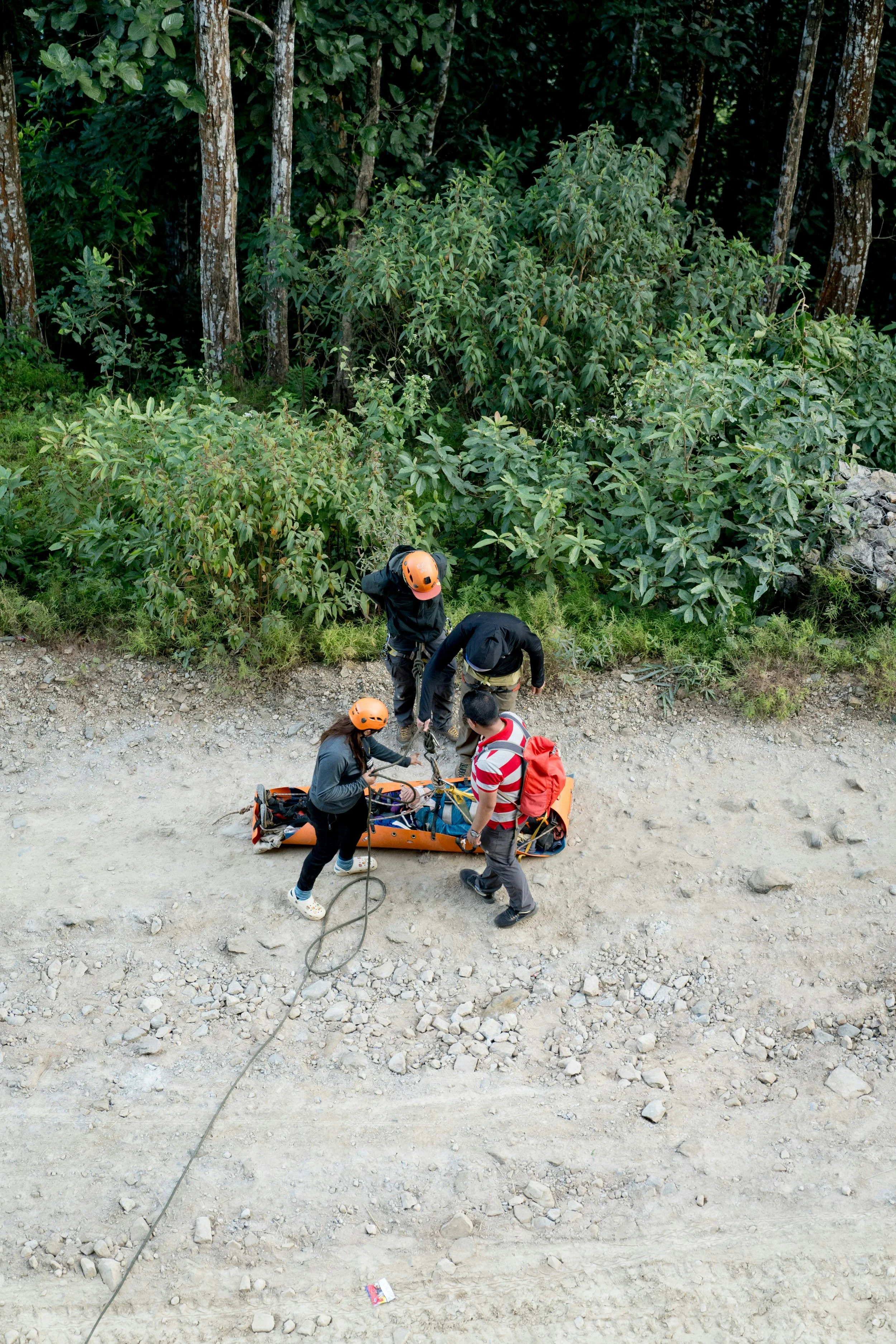 Group of four rescue workers preparing a stretcher with one person on a dirt road surrounded by greenery and trees.