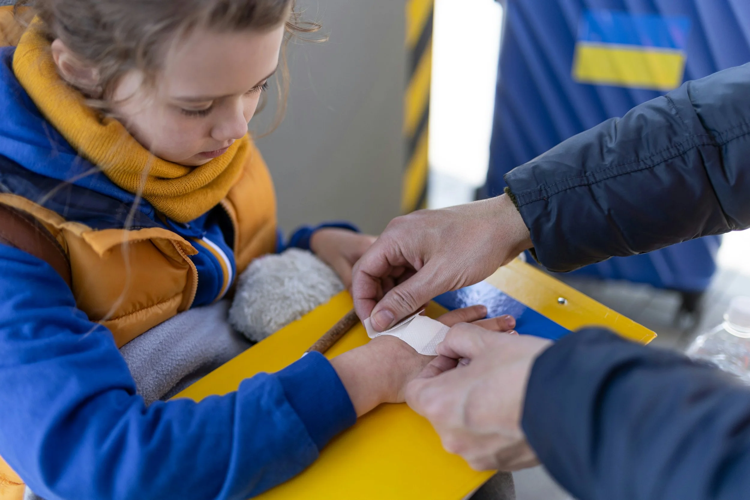 A young girl getting a medical bandage applied on her hand by a person. The girl wears a blue jacket with a yellow collar. The person applying the bandage wears a dark jacket. The scene appears to be indoors or in an emergency setting with yellow and blue background elements.