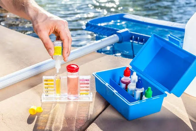 Water testing kits and chemicals on a pool deck, with test tubes and a blue storage container for supplies.
