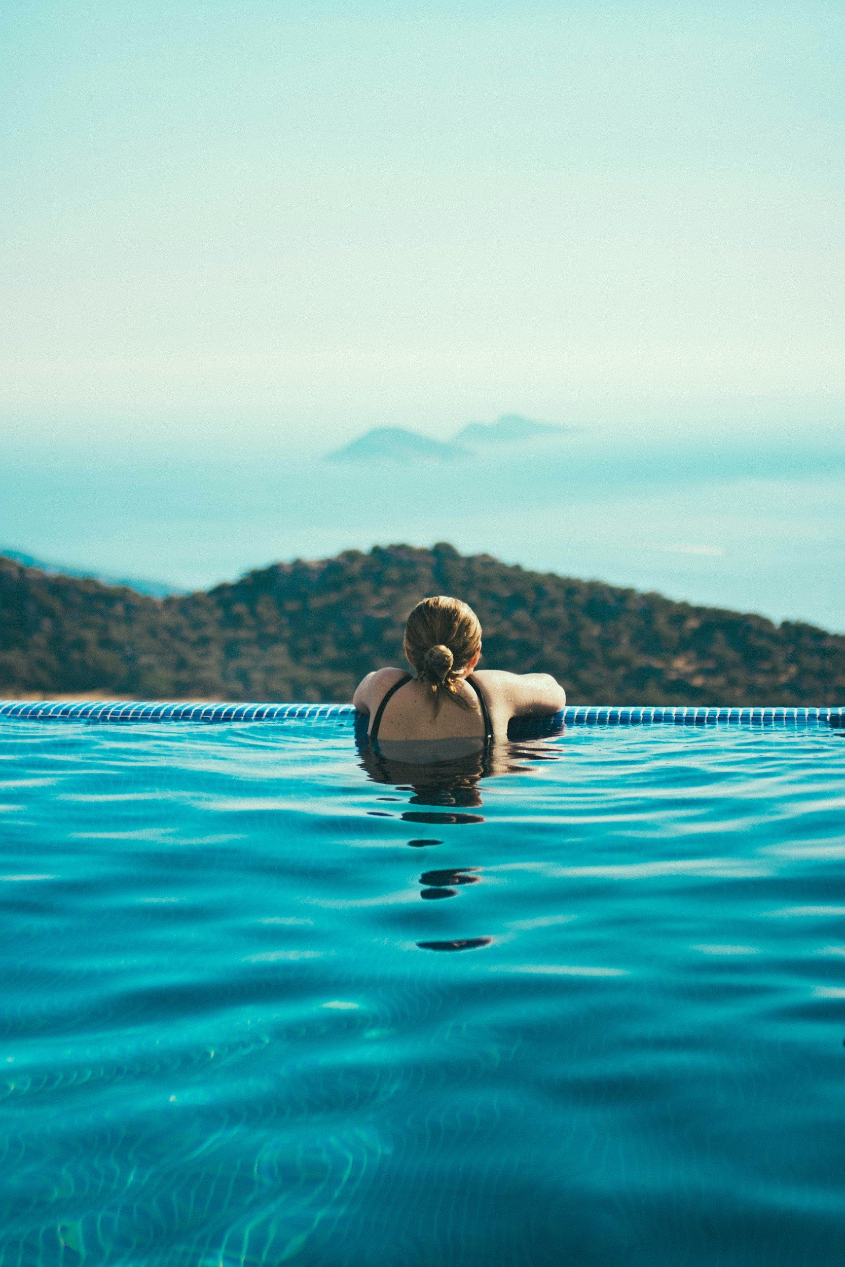 A woman with blond hair in a bun, wearing a black swimsuit, relaxing in an infinity pool with mountains and clouds in the background.