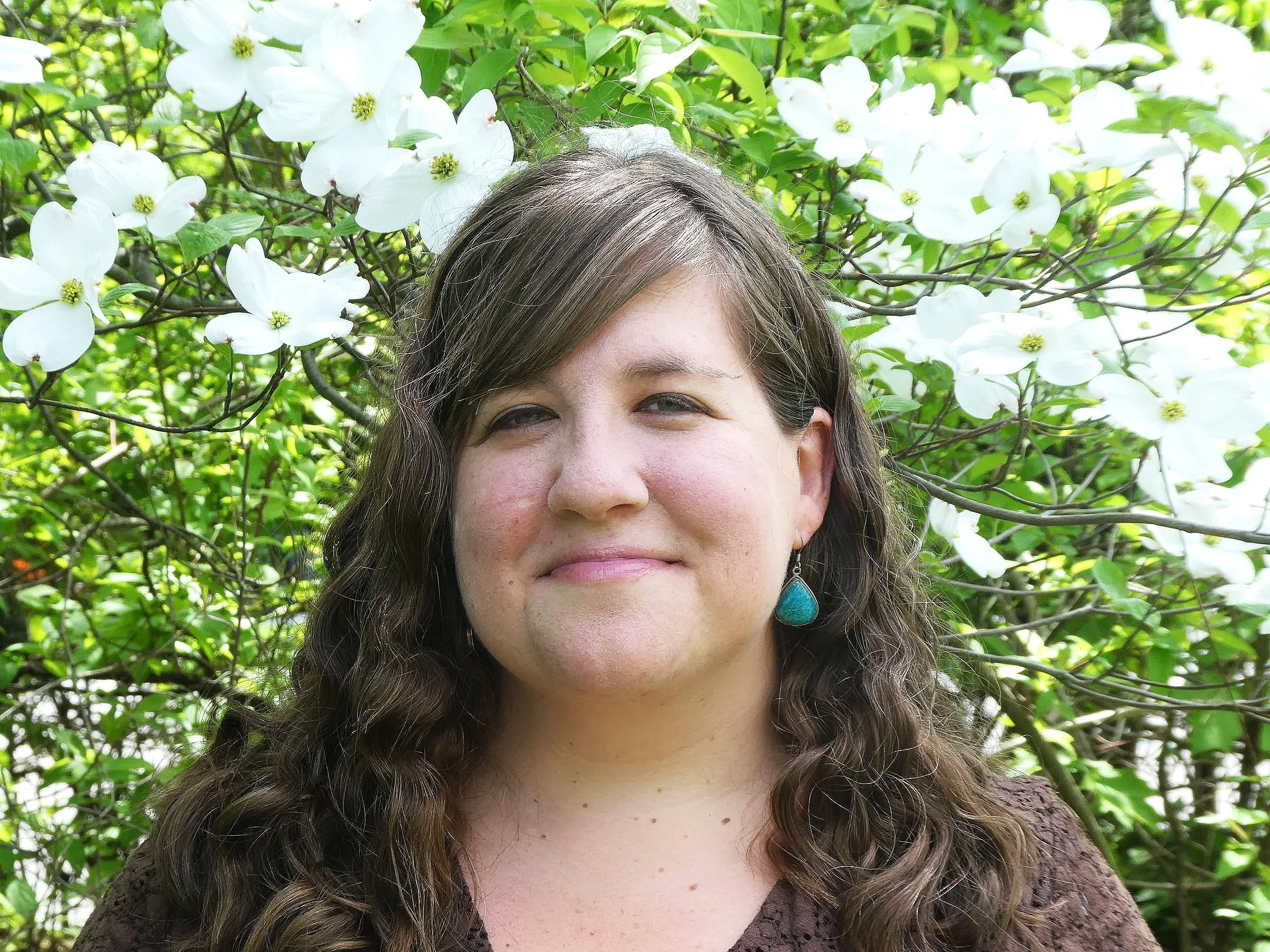 A woman with wavy hair and a turquoise earring smiling outdoors surrounded by white flowering dogwood blossoms and green leaves.