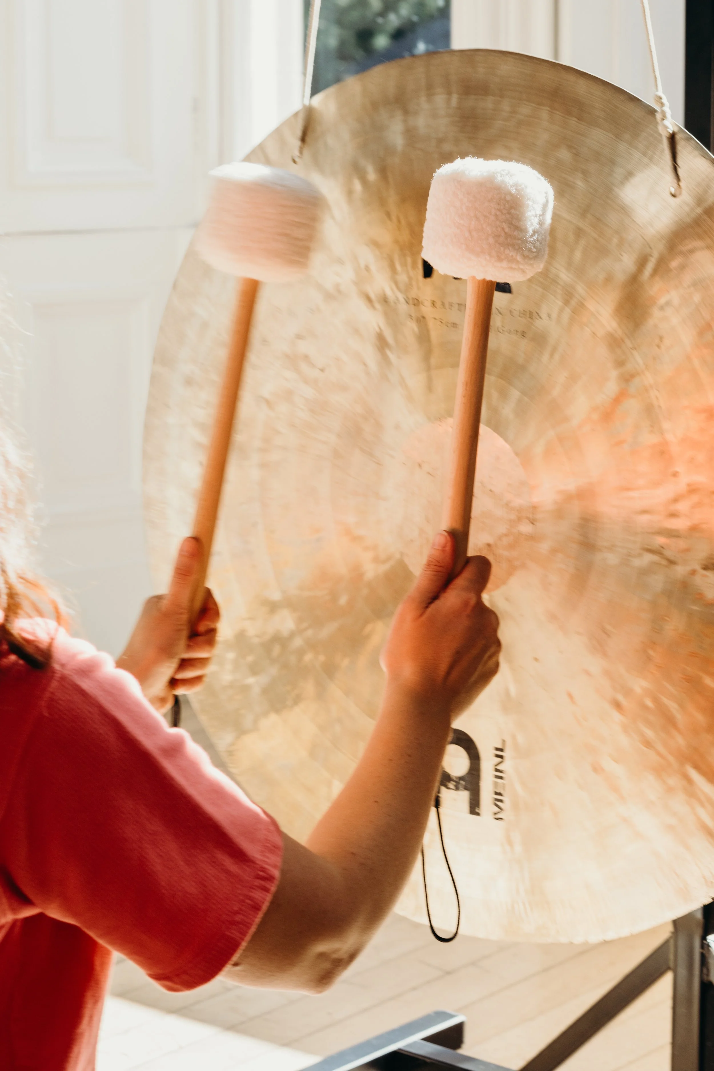 Two hands, one on each side, holding percussion mallets with pink fluffy heads, striking a large bronze gong.