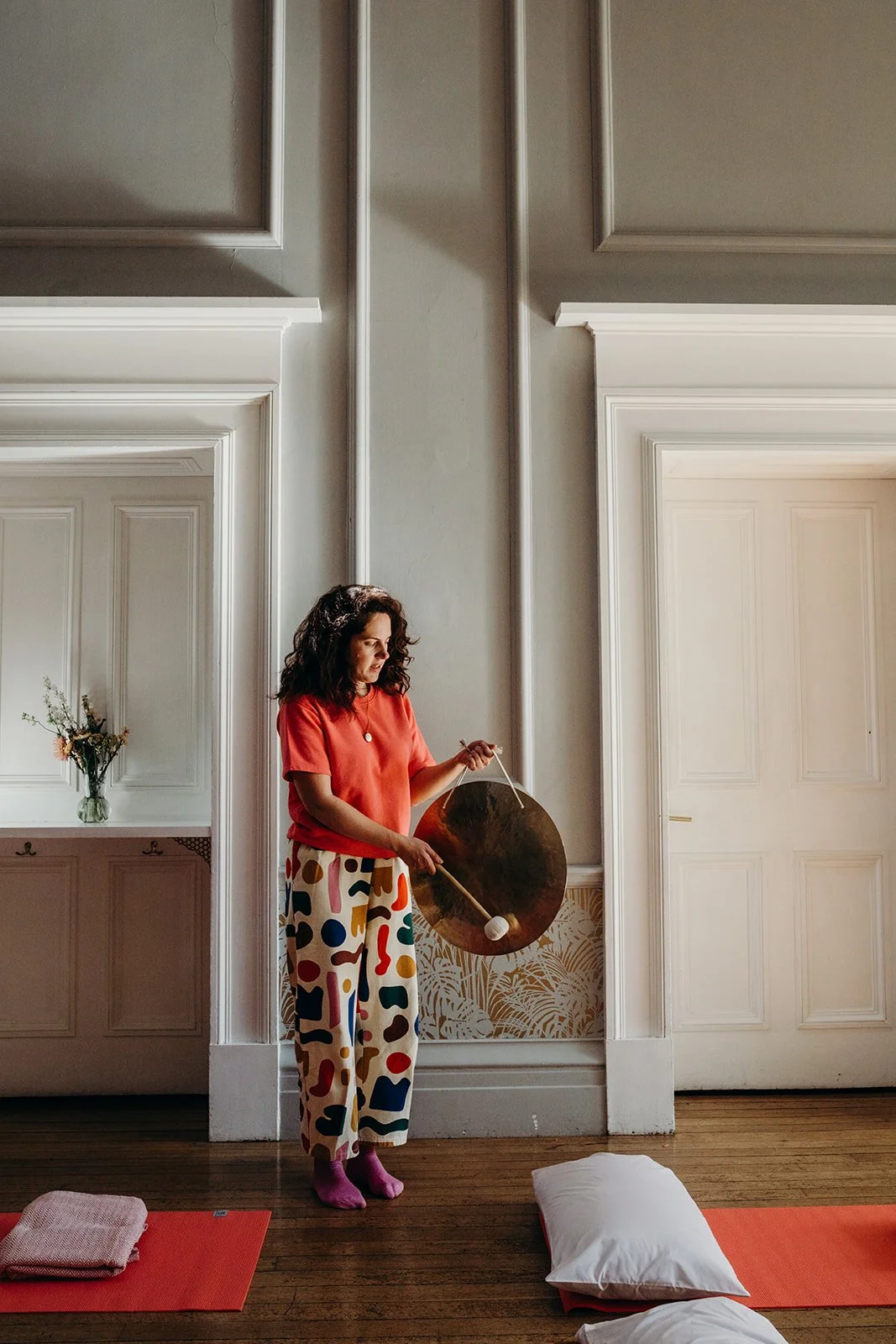 A woman standing in a room with white paneled walls, holding a large gong and a mallet, wearing colorful, patterned pants and pink socks.