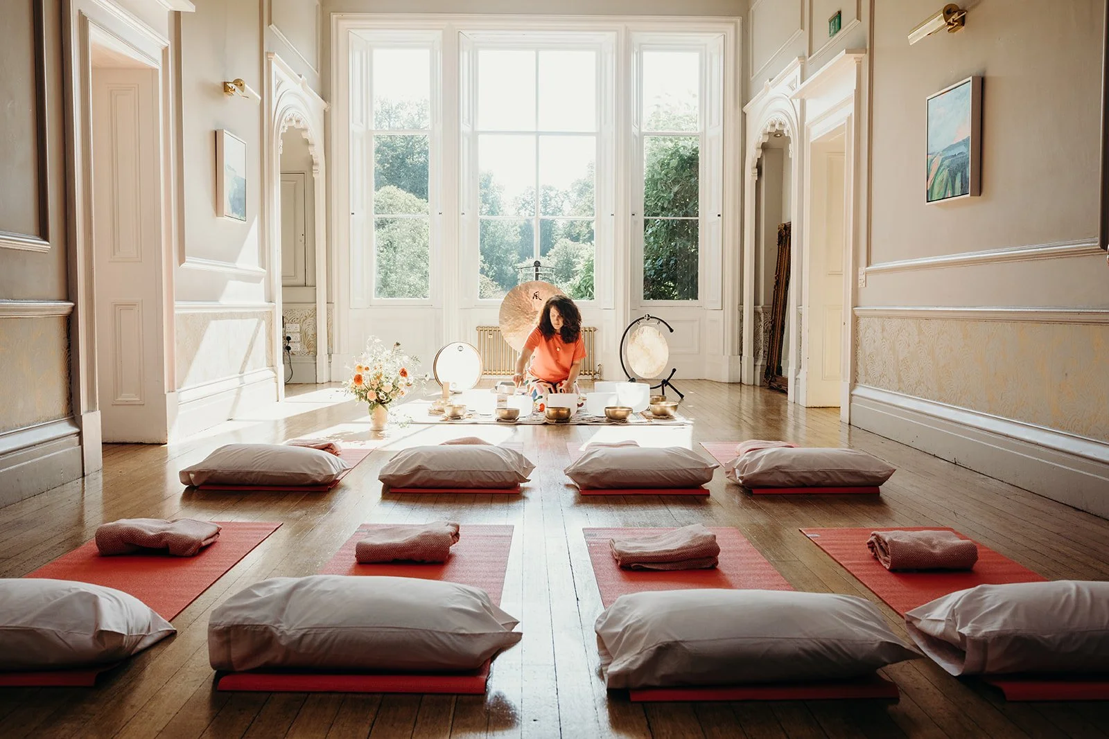 A woman sitting on the floor in a bright, spacious room with large windows, surrounded by yoga mats, pillows, and sound bowls, possibly preparing for a sound healing session.