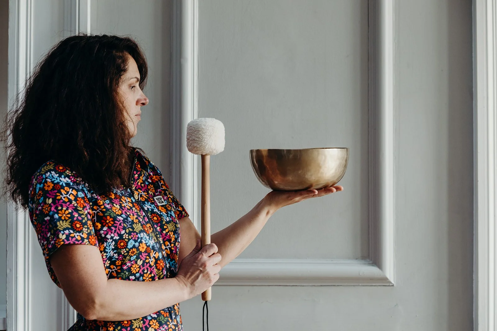 Woman in colorful floral shirt holding a mallet and brass singing bowl with her other hand.