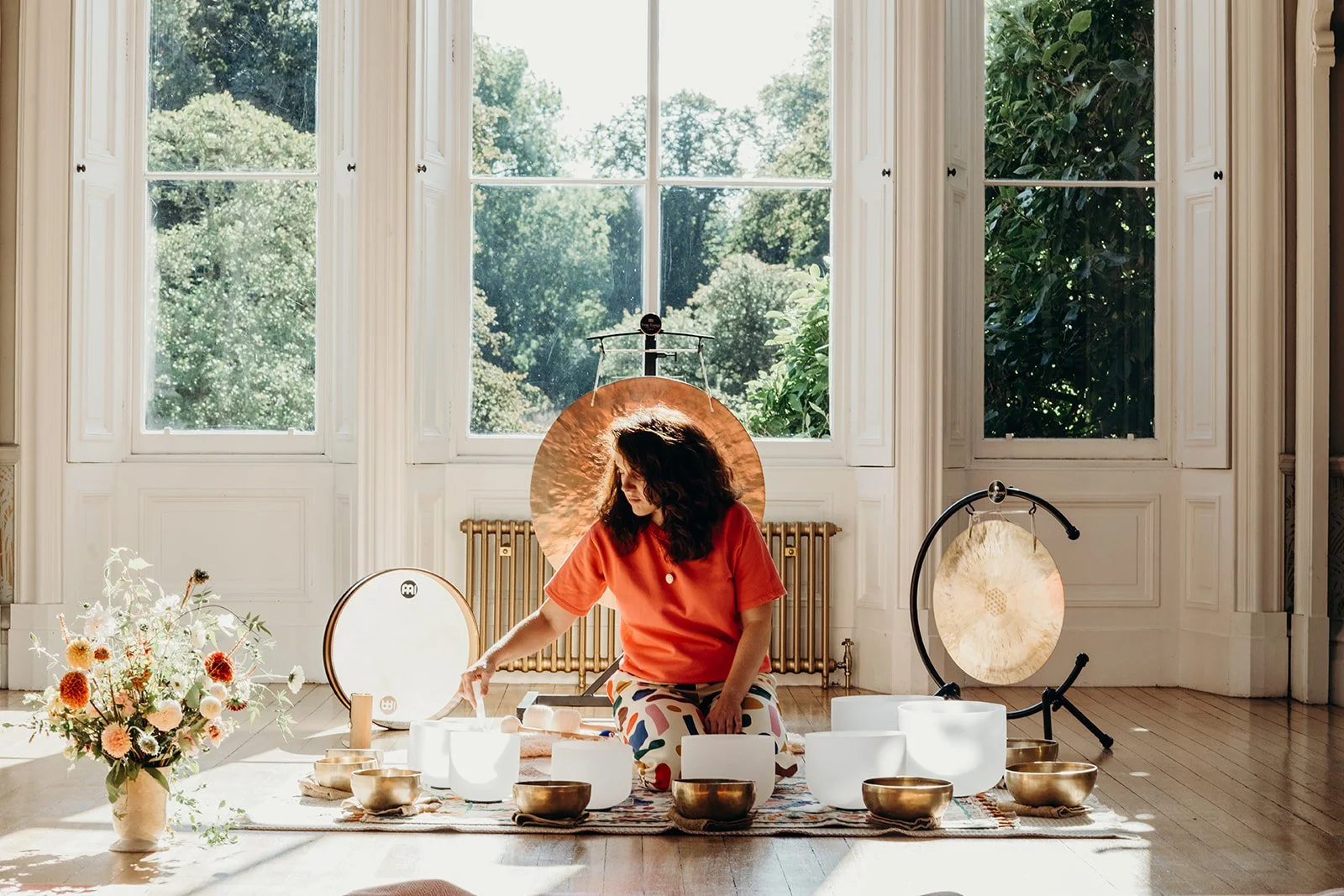 A woman in a red top and patterned pants sitting on the floor surrounded by singing bowls, a gong, and a bouquet of flowers, practicing sound therapy in a sunlit room with large windows and greenery outside.