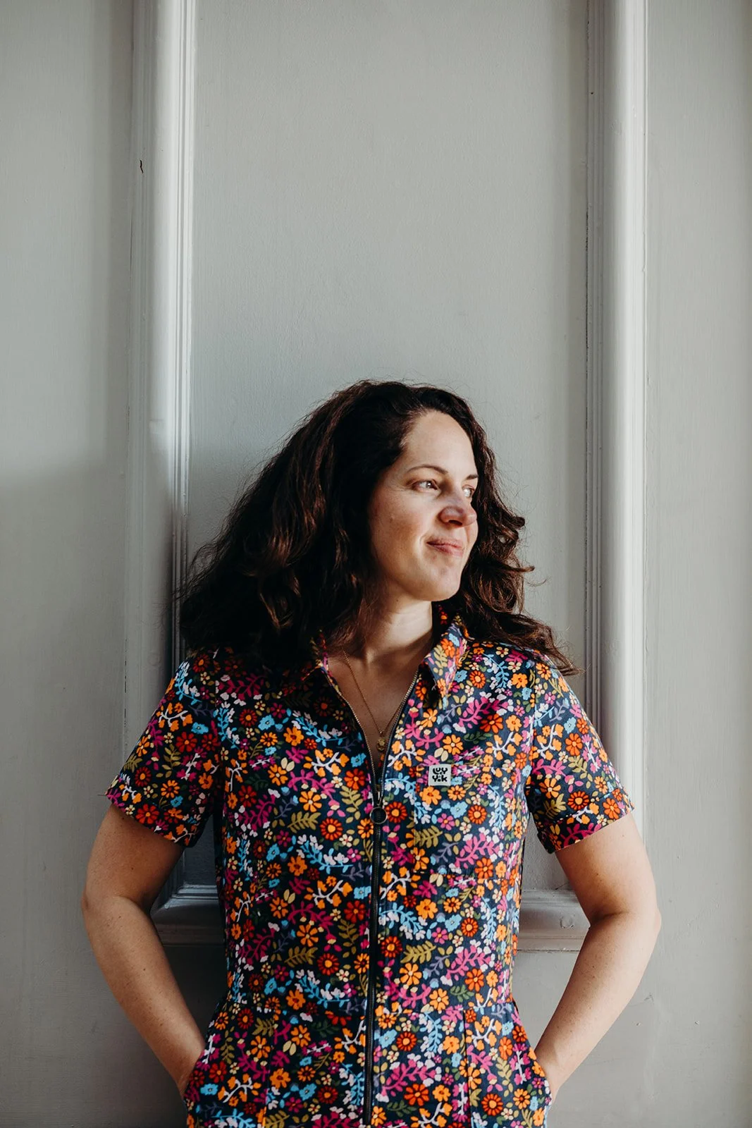 A woman with dark curly hair wearing a colorful floral short-sleeve shirt stands indoors against a white wall with decorative molding, looking off to the side with a slight smile.