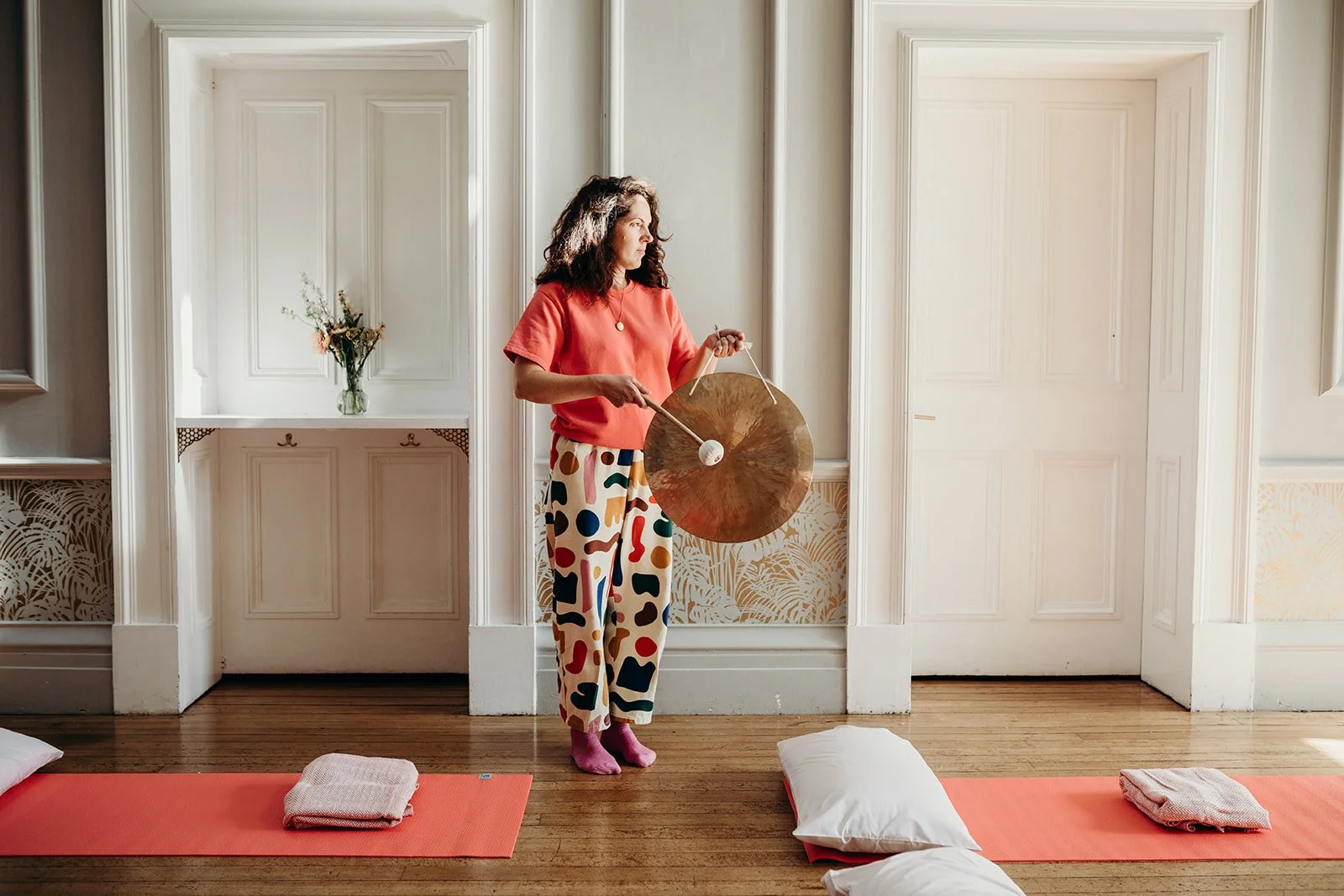 A woman practicing sound therapy with a gong, standing in front of a wall with decorative paneling, surrounded by yoga mats, pillows, and blankets in a bright, airy room.